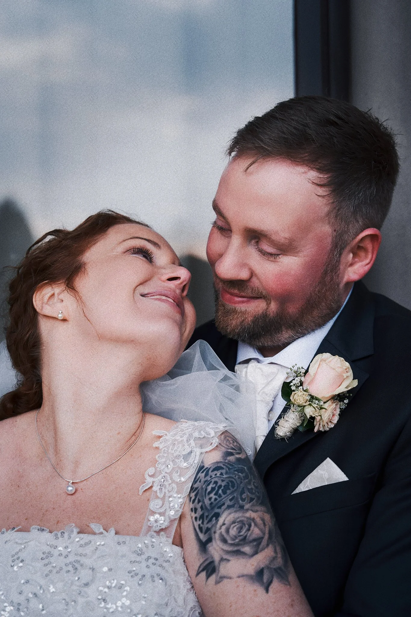 Ein Brautpaar beim Hochzeitsfoto, die Frau trägt ein weißes Brautkleid mit Perlen und Spitze, der Mann trägt einen schwarzen Anzug mit weißem Hemd und Fliege, sie schauen sich liebevoll an, im Hintergrund der Himmel.