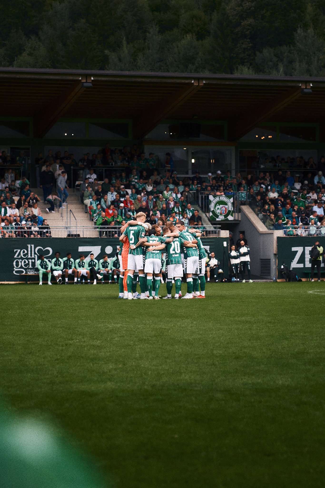 Fußballspieler in grünen Trikots umarmen sich auf dem Spielfeld, im Hintergrund sind Tribünen mit Zuschauern, bei Dämmerung.