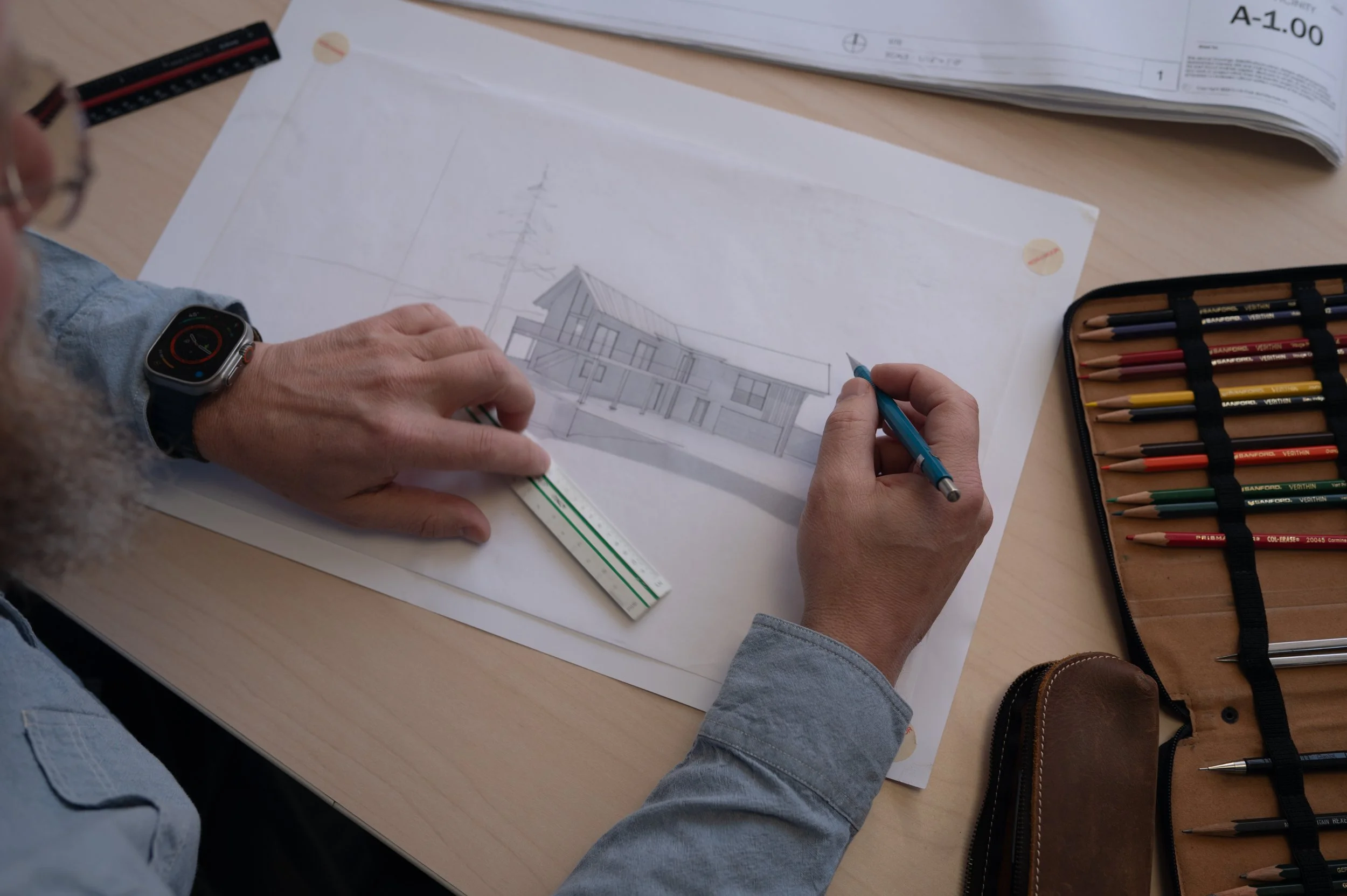 A person working on architectural plans with a scale ruler on a light wood table. The person wears a smartwatch and a light blue shirt. There is a set of colored pencils in a black case and a large stack of papers with building elevations nearby.