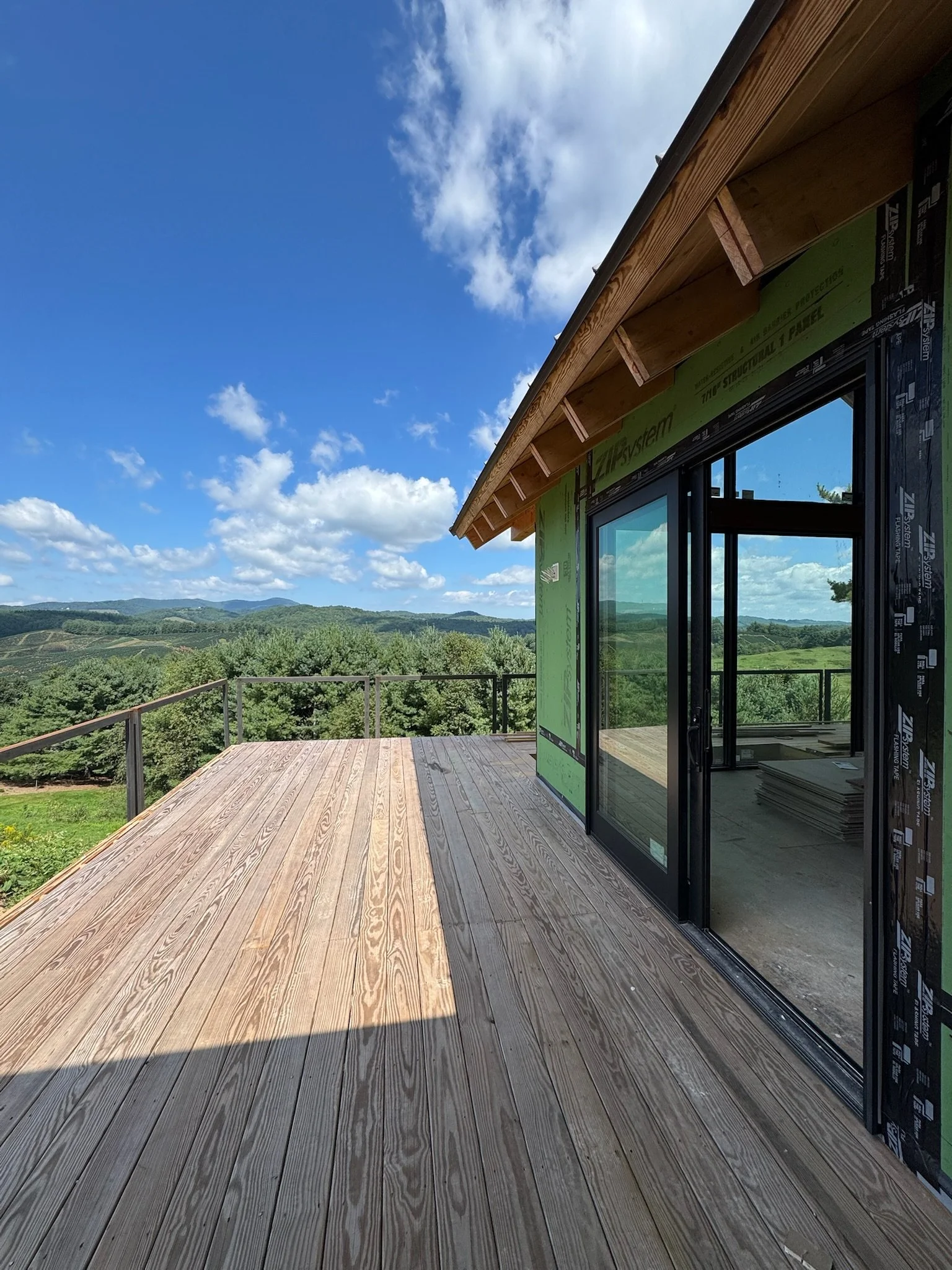 Partial view of a house under construction with a wooden deck, black-framed glass doors, and a landscape of rolling hills and a blue sky with scattered clouds in the background.