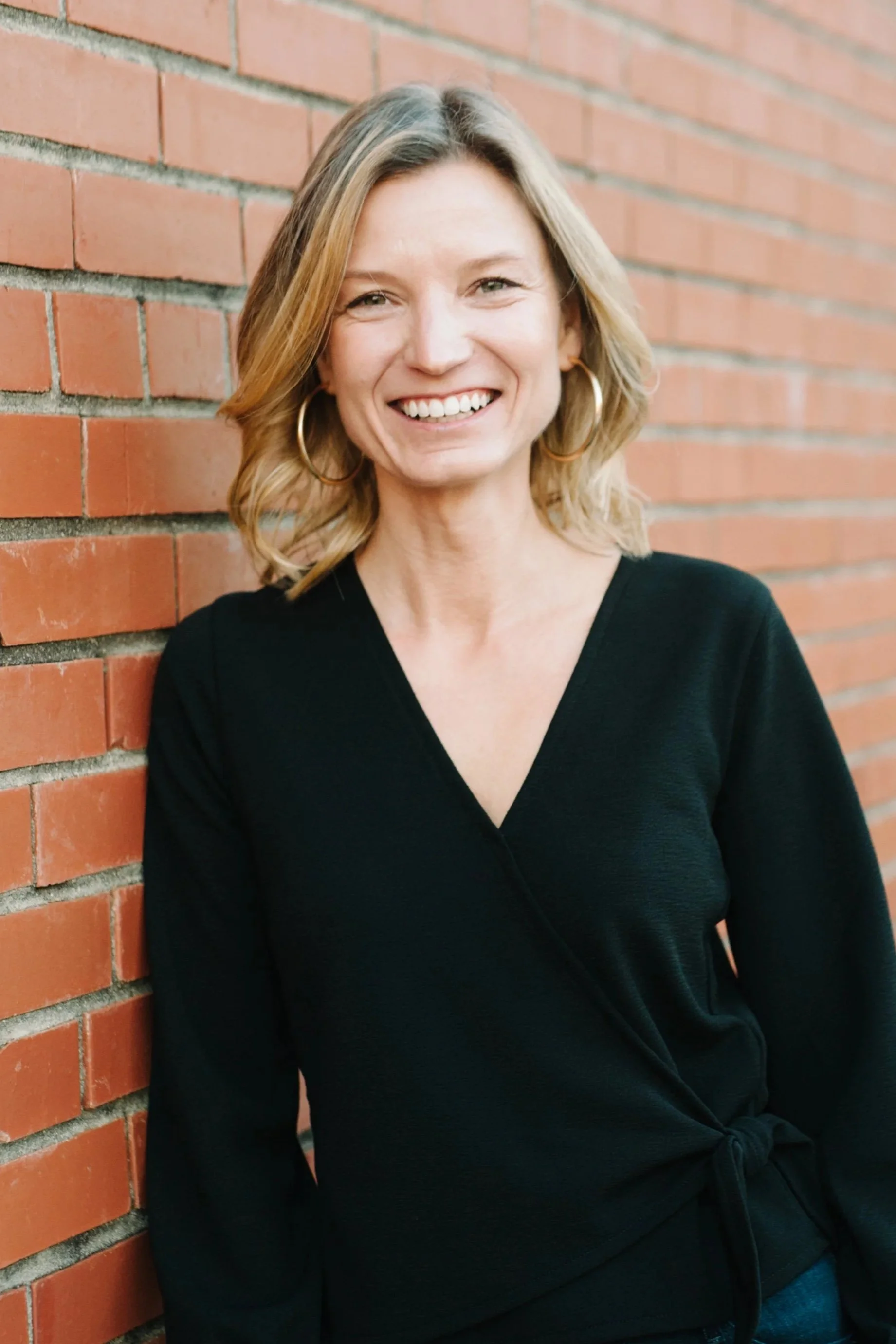 A woman with shoulder-length blonde hair, hoop earrings, and a black top smiling while leaning against a brick wall.