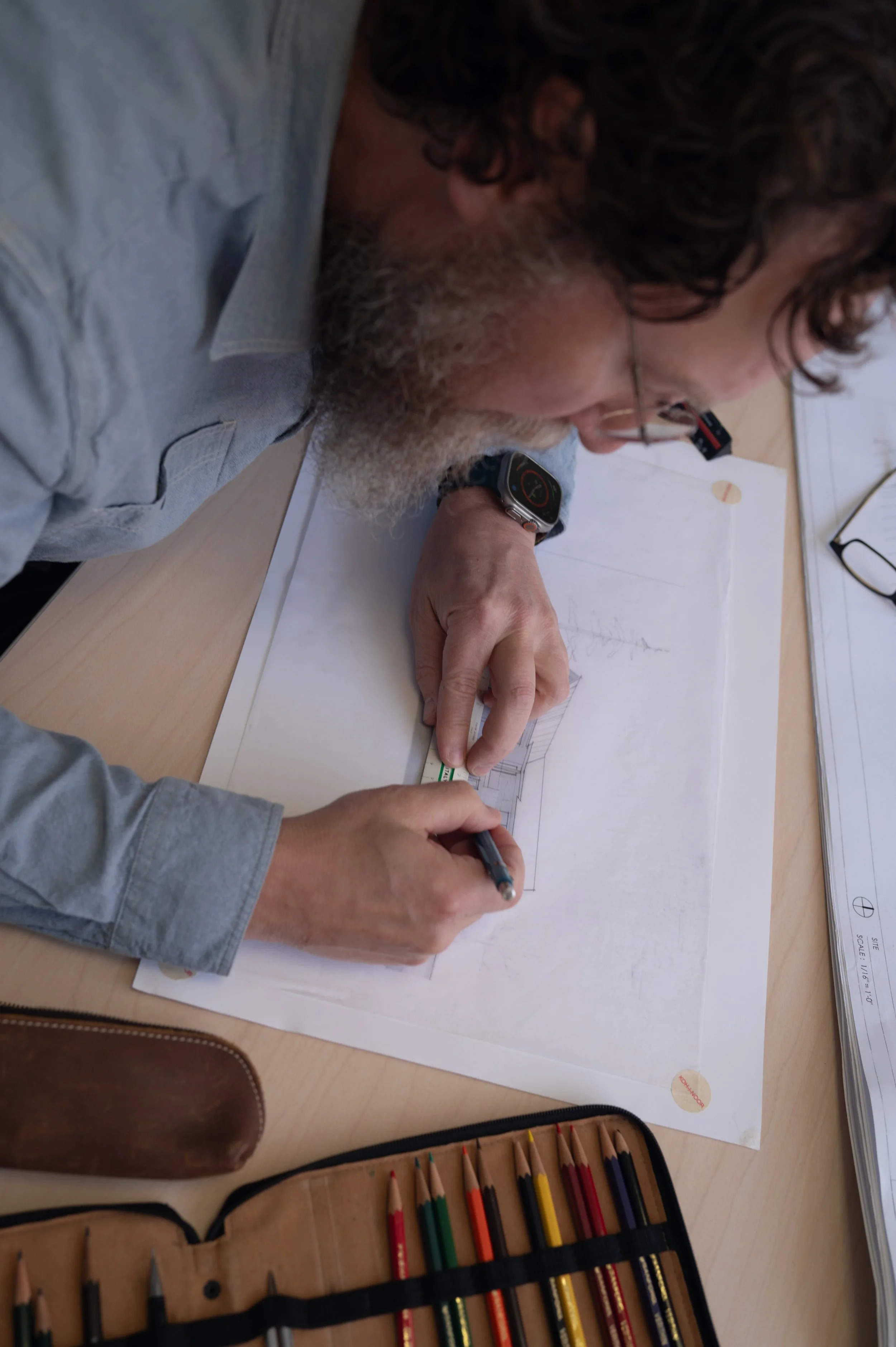 A man with curly hair and glasses is leaning over a table, drawing architectural plans on a sheet of paper. There are colored pencils in a case and a pair of glasses on the table.