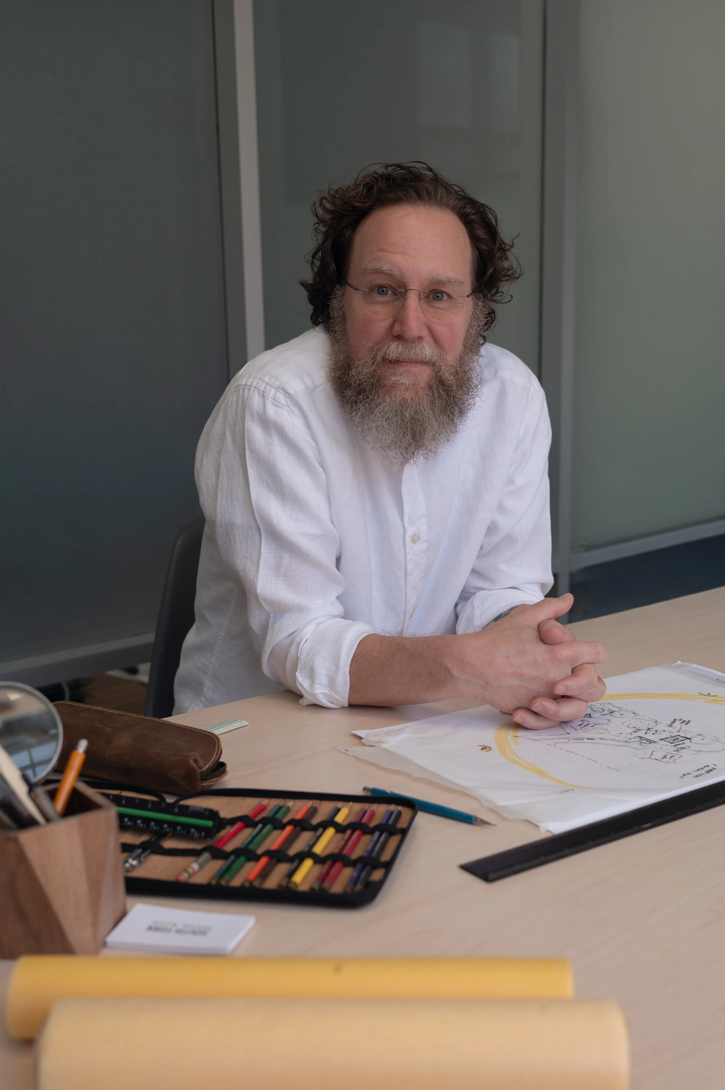A man with glasses and a beard sitting at a desk, smiling at the camera. The desk has art supplies, including colorful pastels, pens, and papers with drawings.