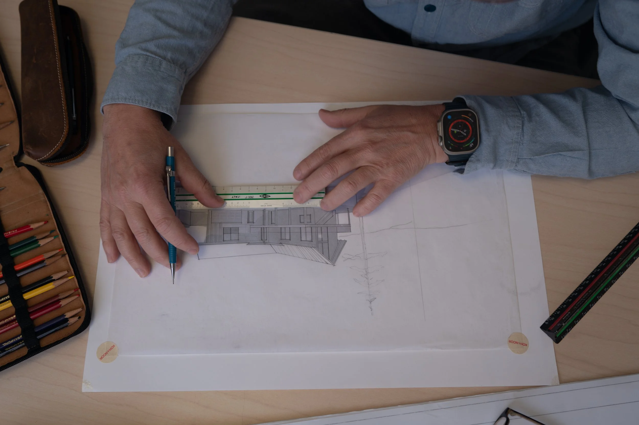 A person wearing a smartwatch is holding a blue pen and working on architectural drawings of a building on a large sheet of paper.