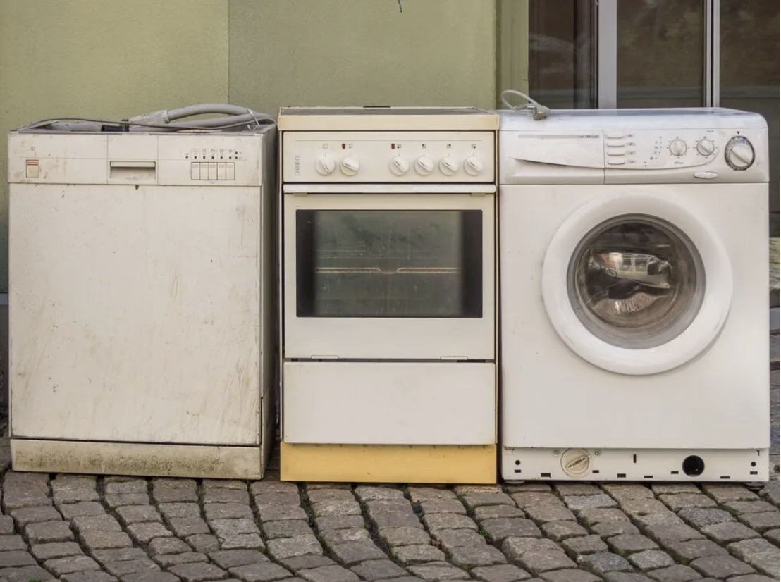 Three old appliances, a dishwasher, a stove, and a washing machine, placed side by side on a cobblestone surface outside against a green wall.