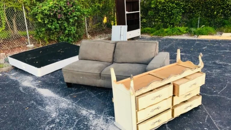Outdoor scene with discarded furniture on a paved surface, including a gray sofa, a broken bed frame, and a vintage dresser with drawers