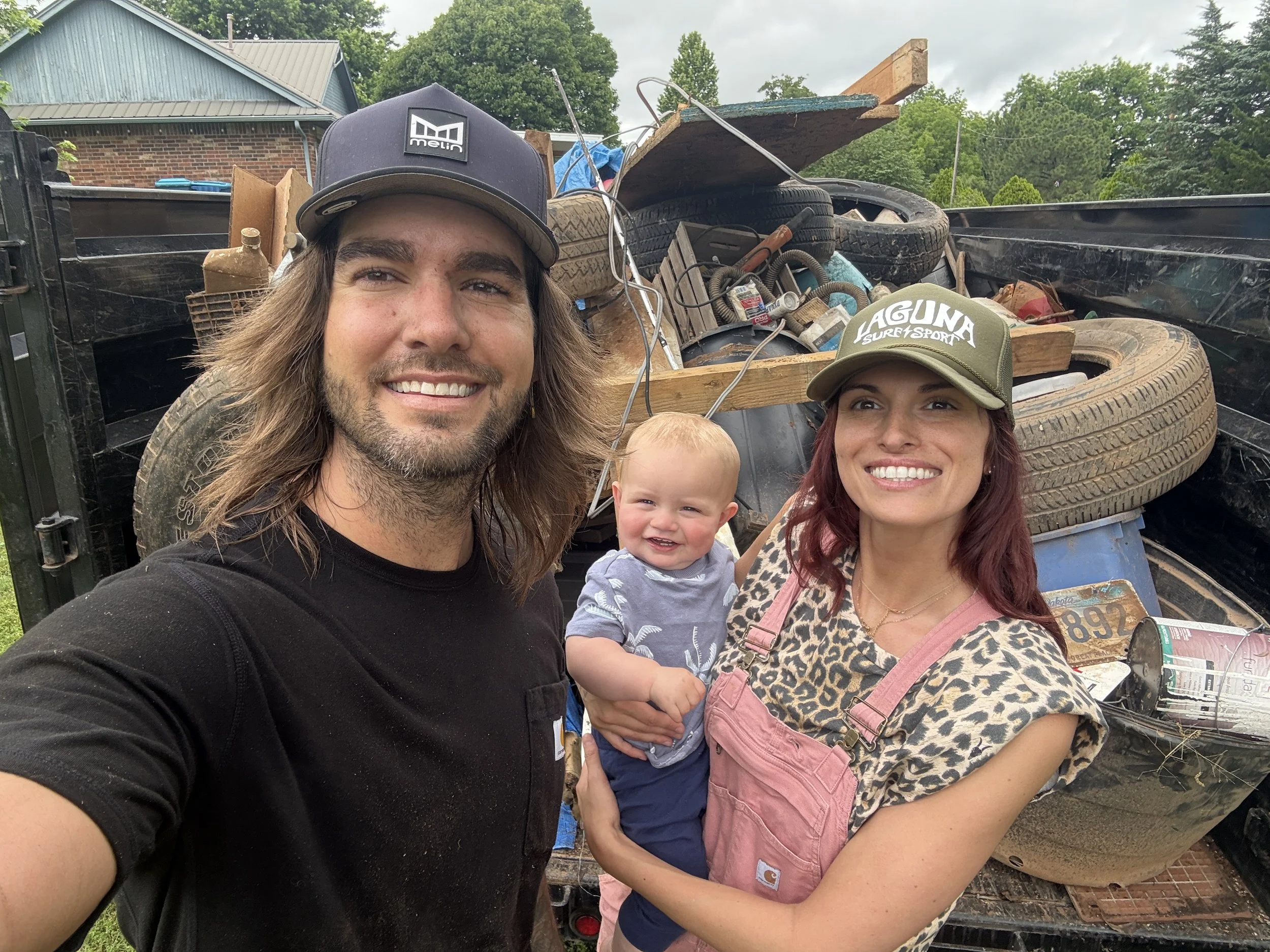 A smiling man with shoulder-length hair, a woman with red hair, and a toddler, all smiling, taking a selfie in front of a truck bed filled with various clutter and old tires, outdoors with trees and buildings in the background.