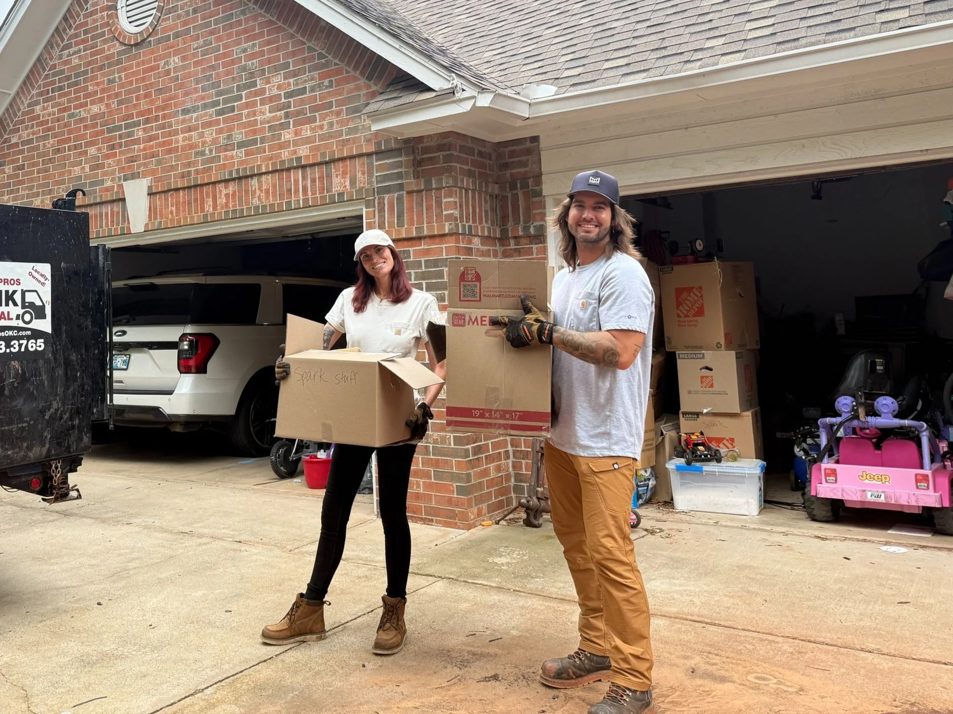 Two people standing outside a garage, holding cardboard boxes, smiling. The woman has red hair and wears a white shirt, black pants, and tan work boots. The man has long hair, wears a hat, gray shirt, tan pants, and gloves. The garage contains boxes, toys, and a small pink toy vehicle.