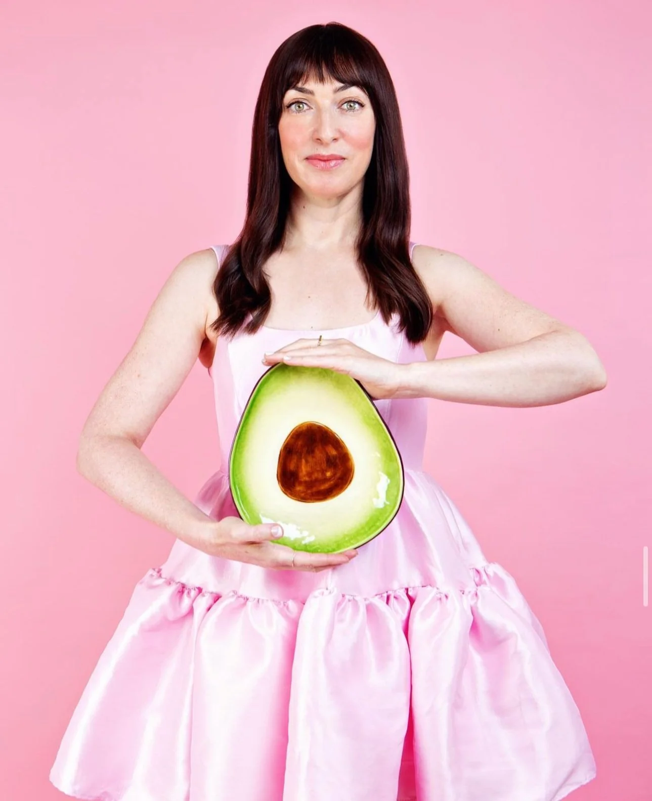 Woman with dark brown hair wearing a pink dress, holding a large avocado with the seed visible in front of a pink background.