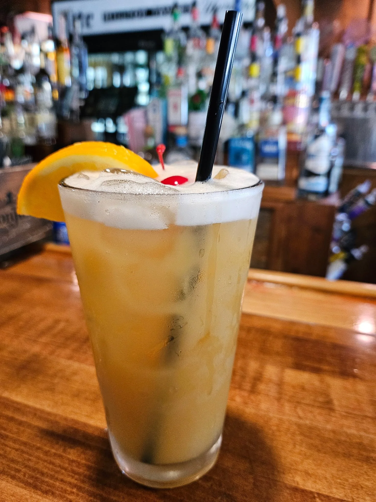 A tall glass of a light-colored cocktail garnished with a lemon wedge, a maraschino cherry, and a black straw, on a wooden bar counter with a blurred bar background.