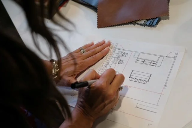 A person drawing architectural or interior design plans on paper with a pen, with fabric samples on the table.