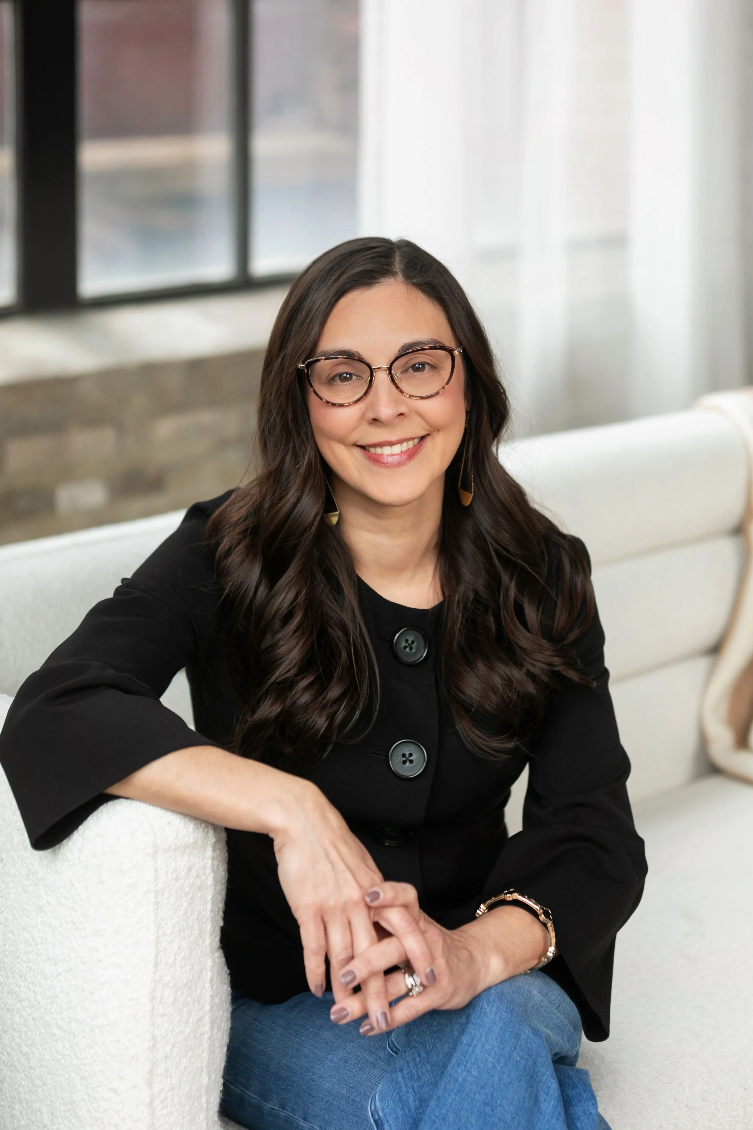 A woman with long dark wavy hair wearing glasses, a black top with large buttons, and jeans, sitting on a white sofa near a window with sheer curtains, smiling at the camera.