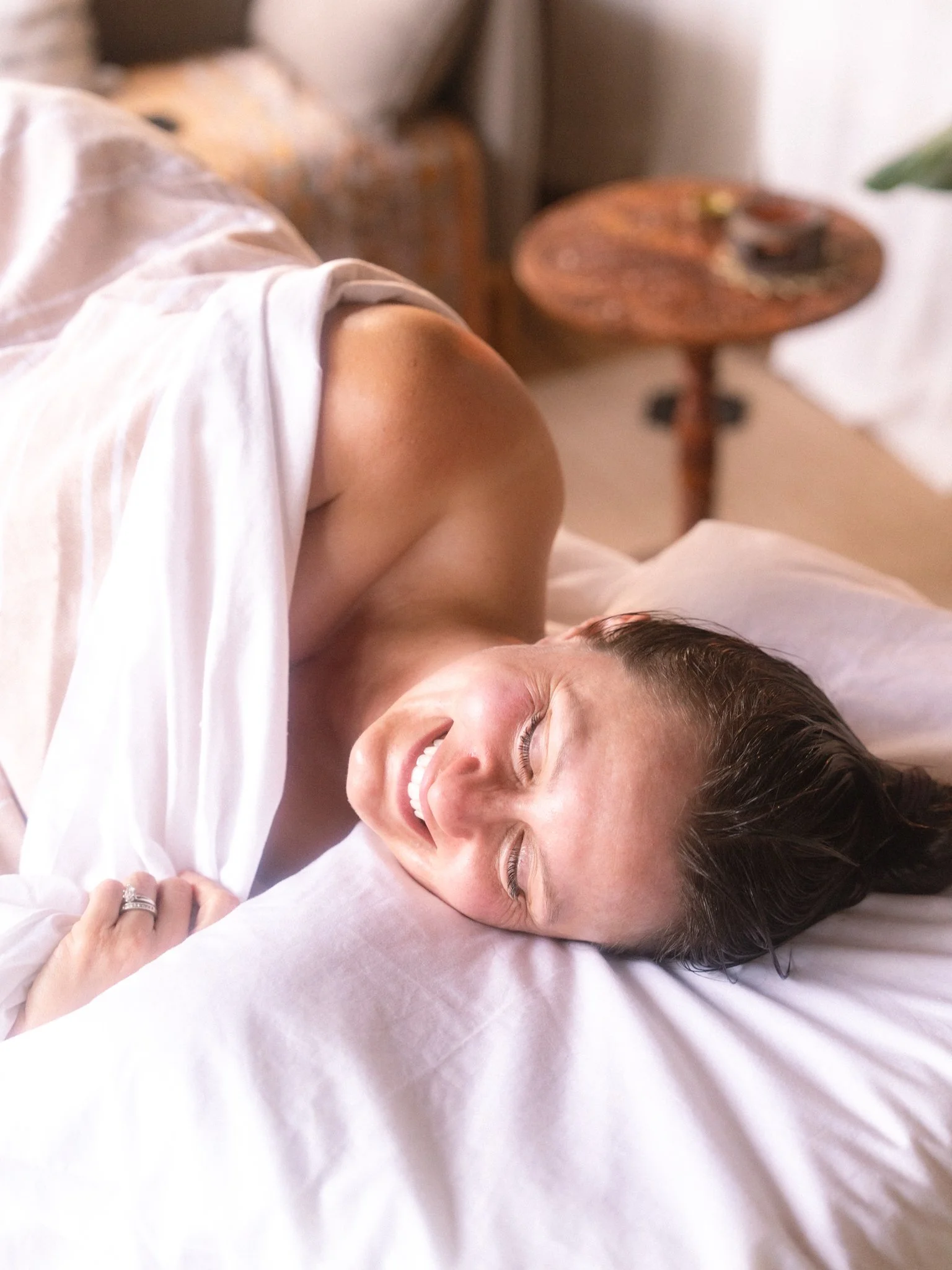 A woman lying on a bed, smiling, with her eyes closed, wearing a white shirt, with a bed pillow behind her head, and a round wooden table in the background.