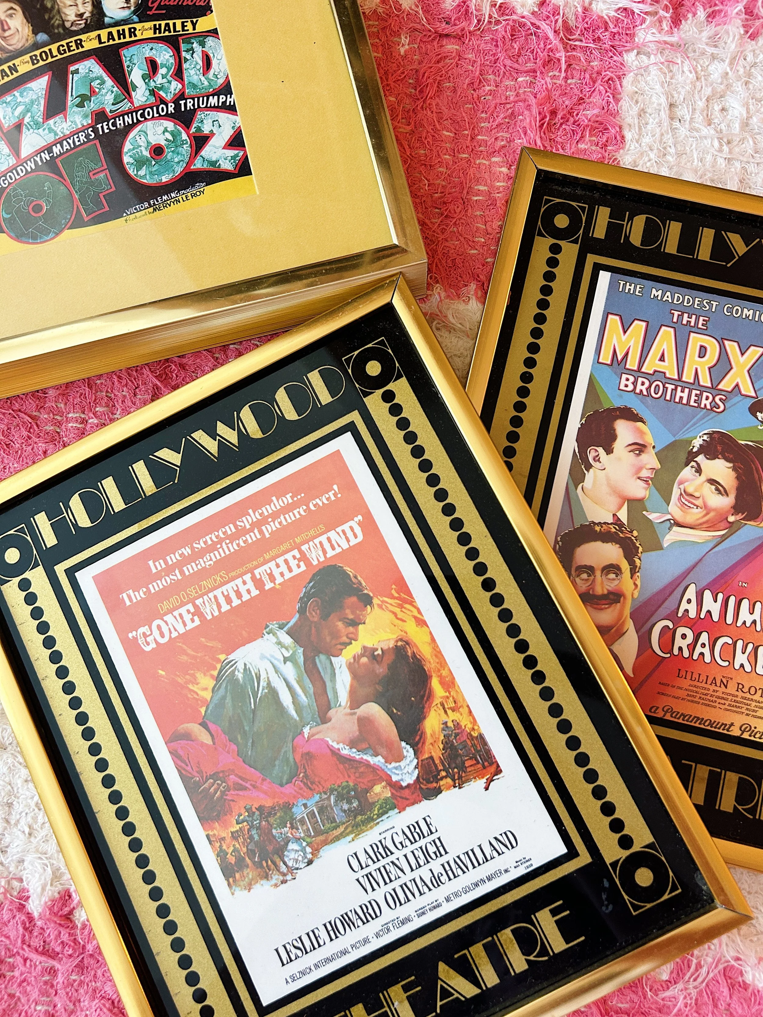 Vintage movie posters in gold frames on a pink textured surface. The prominent poster is for the film 'Gone with the Wind,' featuring Clark Gable and Vivien Leigh, with the title in bold red letters and a romantic scene of the two characters. 