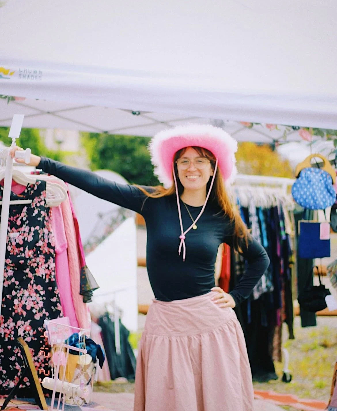 A smiling woman wearing a large pink and white fuzzy hat, glasses, a black long-sleeve shirt, and a light pink skirt, standing under a white outdoor canopy at a market stall with clothing and accessories.