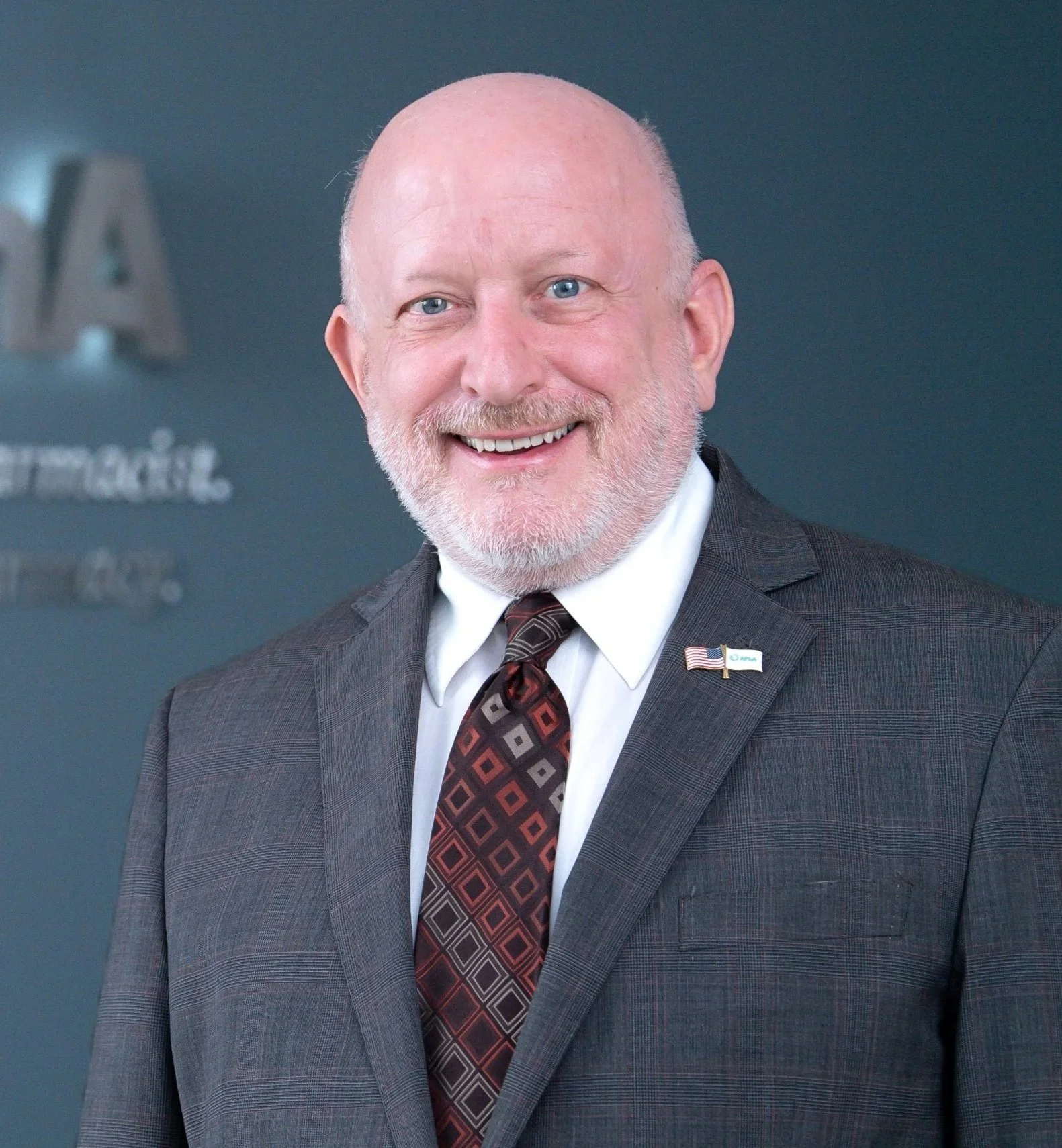 A smiling, bald, older man with a white beard, wearing a suit and tie, standing in front of a dark blue background with blurred signage that includes a logo and American flag pin on his lapel.