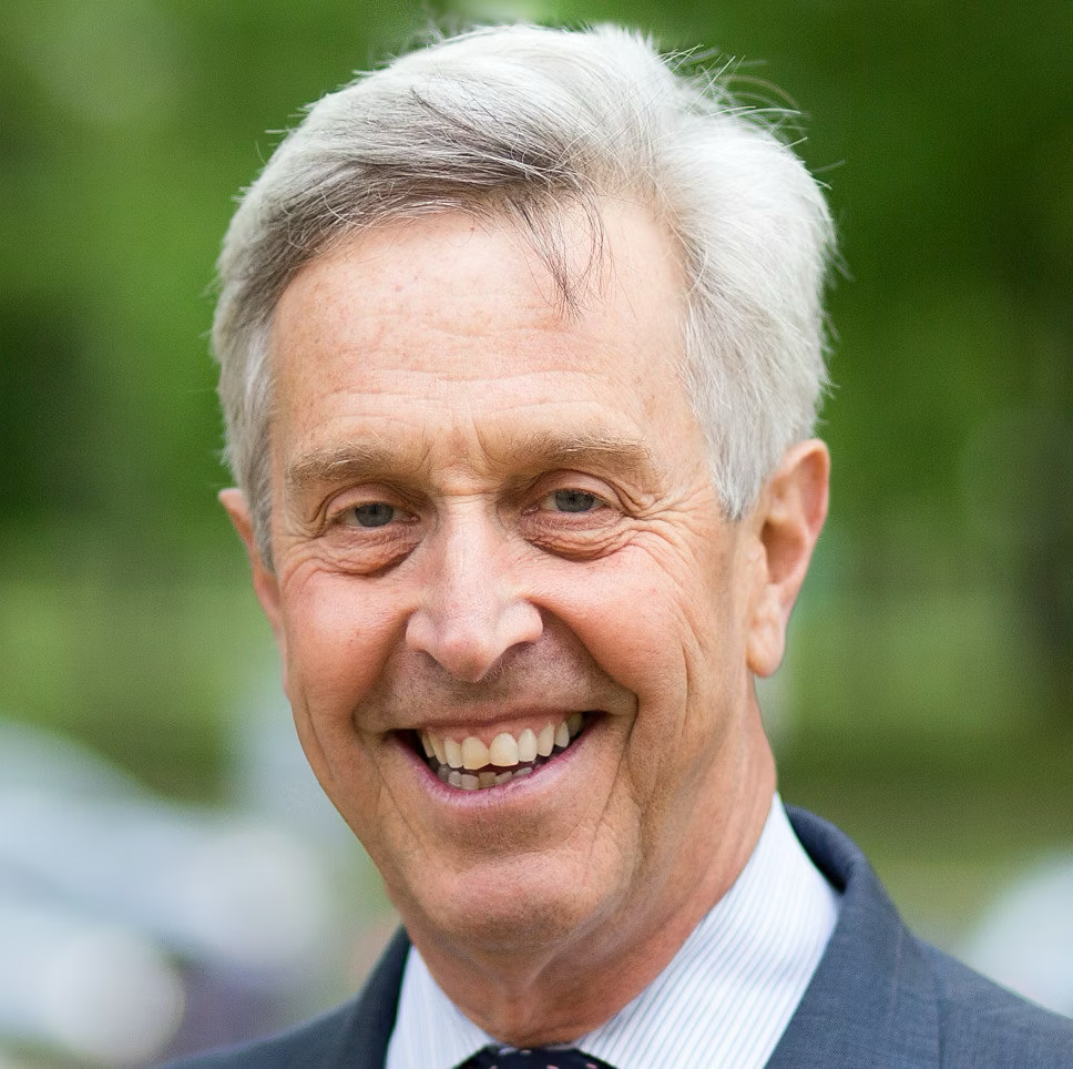 Smiling elderly man with gray hair wearing a suit and tie, outdoors with green blurred background.