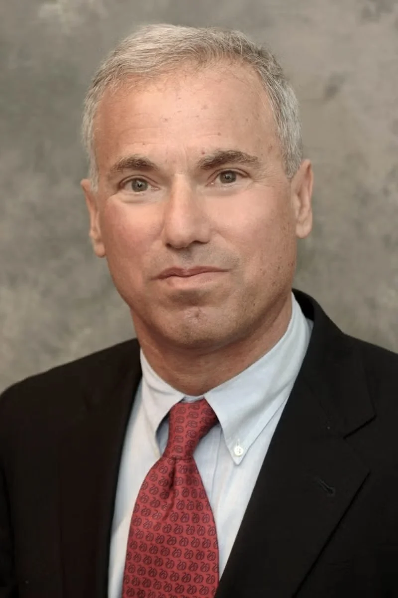 A portrait of a middle-aged man with short gray hair, fair skin, wearing a black blazer, light blue dress shirt, and red patterned tie, against a gray textured background.