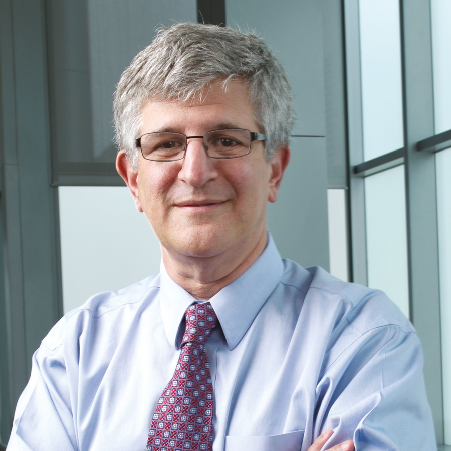 Portrait of a middle-aged man with gray hair, glasses, wearing a light blue dress shirt and a patterned tie, standing inside a modern office with large glass windows.