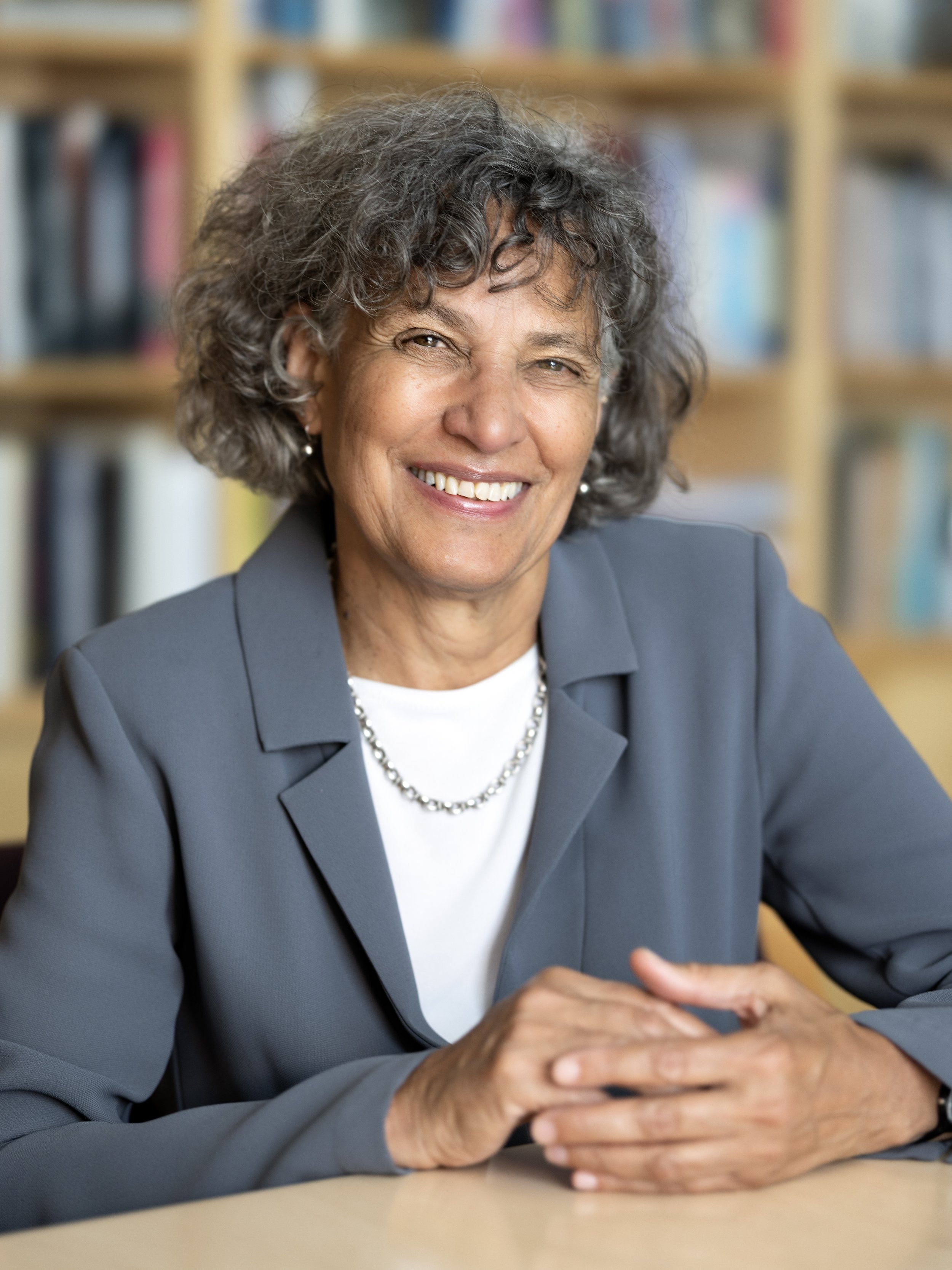 A smiling mature woman with curly gray hair, wearing a gray blazer, white shirt, and pearl necklace, sitting at a table with bookshelves in the background.