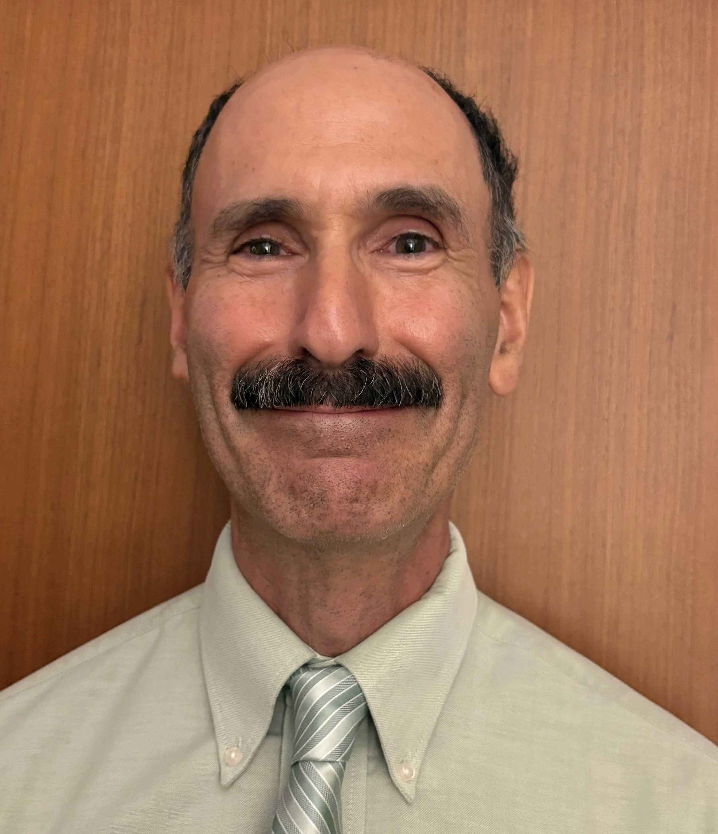 A middle-aged man with a bald head and a mustache, wearing a light-colored dress shirt with a striped tie, standing in front of a wooden background.