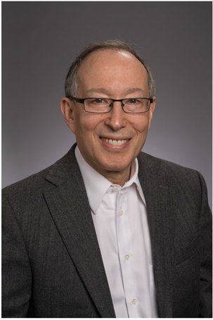 Portrait of a middle-aged man smiling, wearing glasses, a white shirt, and a gray blazer against a gray background.