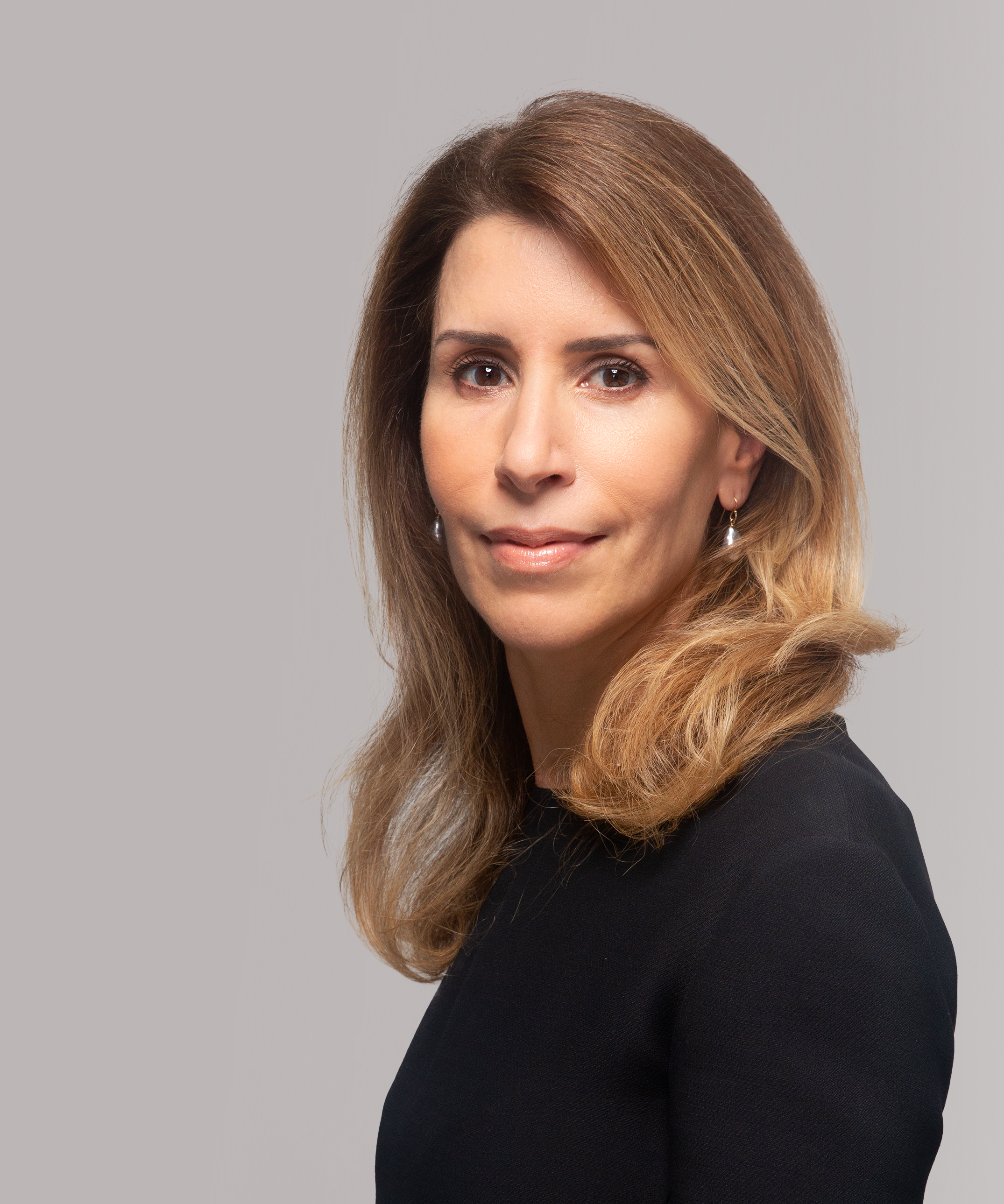 A woman with shoulder-length wavy light brown hair, wearing a black top and pearl earrings, looking at the camera with a slight smile against a gray background.