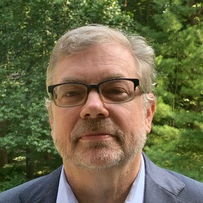 Close-up of a middle-aged man with glasses and a beard, outdoors with green trees in the background.