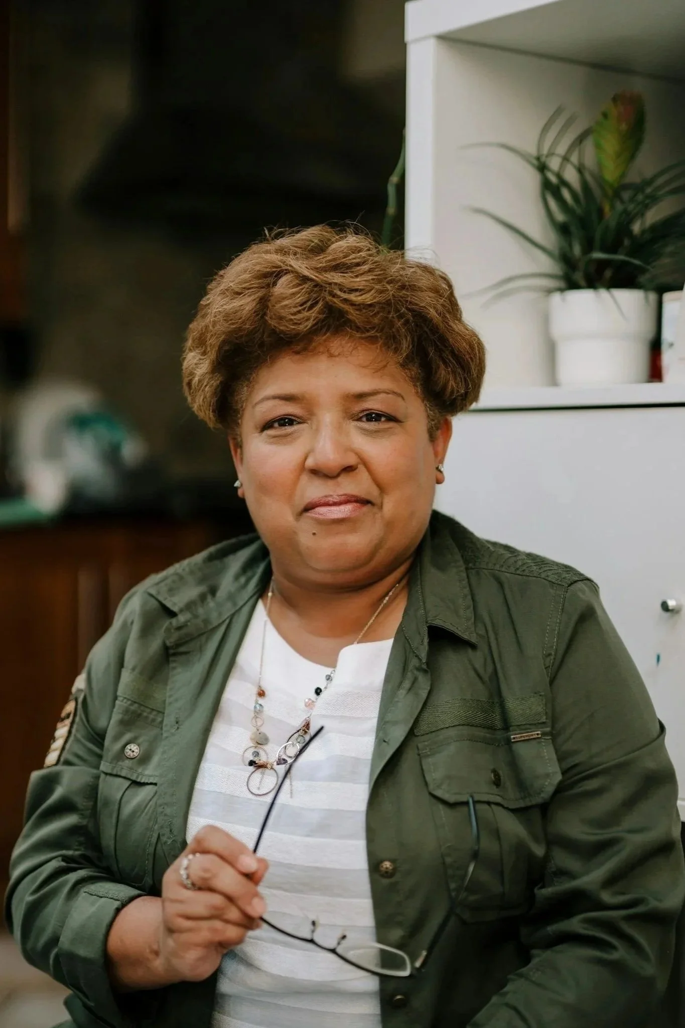 A woman with short, curly brown hair, wearing a green jacket over a white striped shirt, holding glasses in her right hand, sitting indoors with a plant on a white shelf in the background.