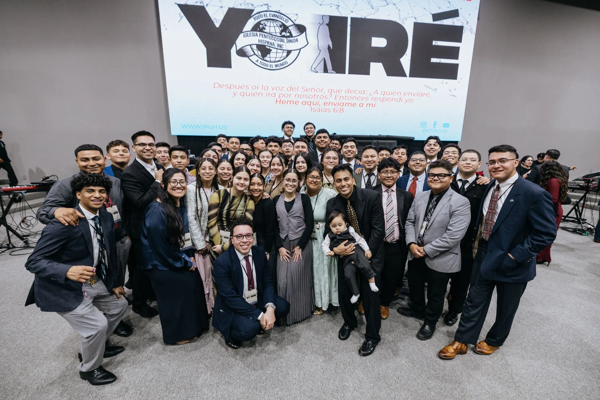 A group of young people and adults dressed in formal attire posing for a photo in front of a large screen displaying a religious message with the logo of an evangelist church.