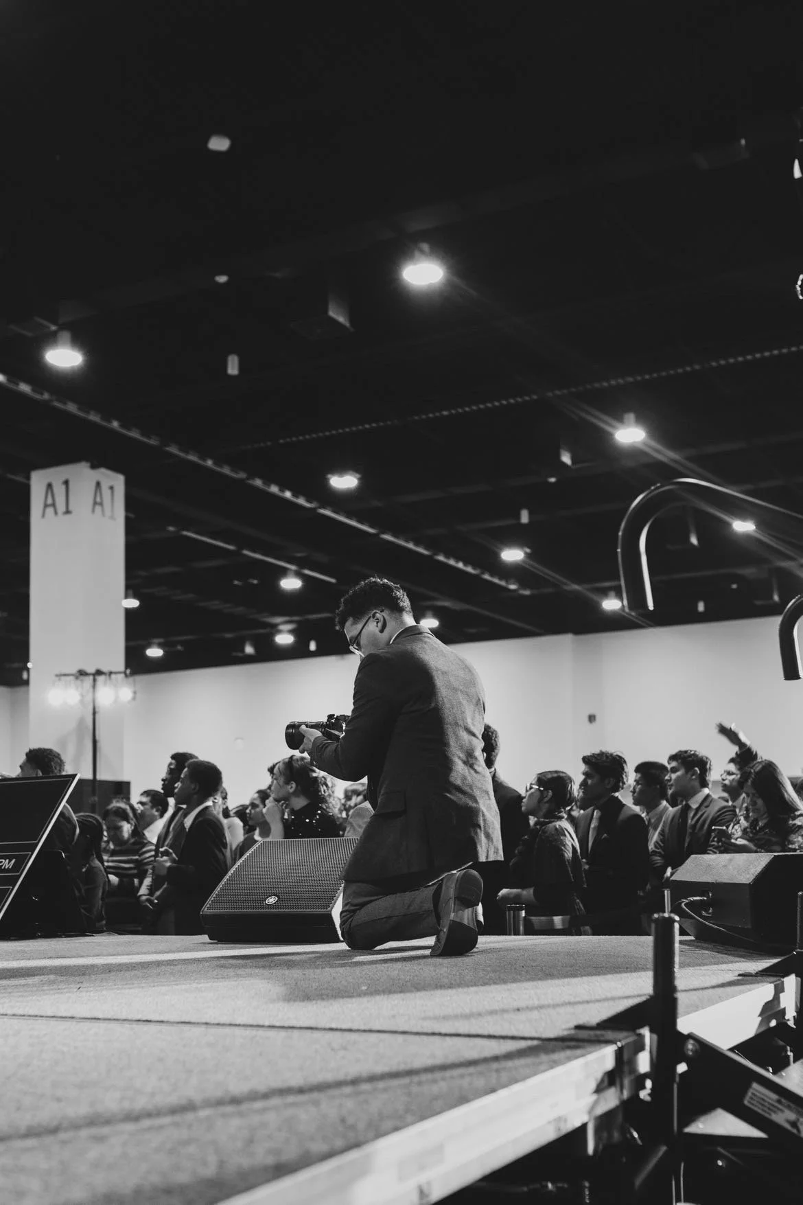 A man in a suit kneeling on a stage with a camera, photographing a seated audience in a conference or event setting.