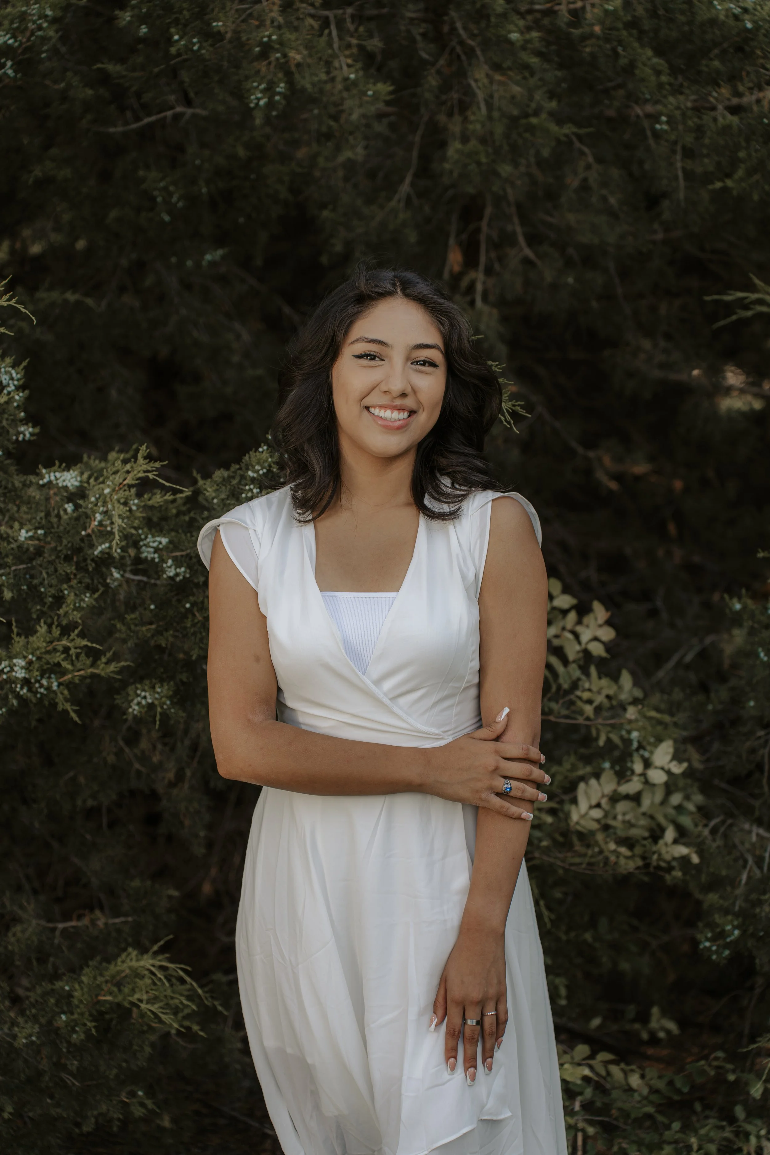 A young woman with dark, wavy hair, wearing a white dress, smiling outdoors with greenery in the background.