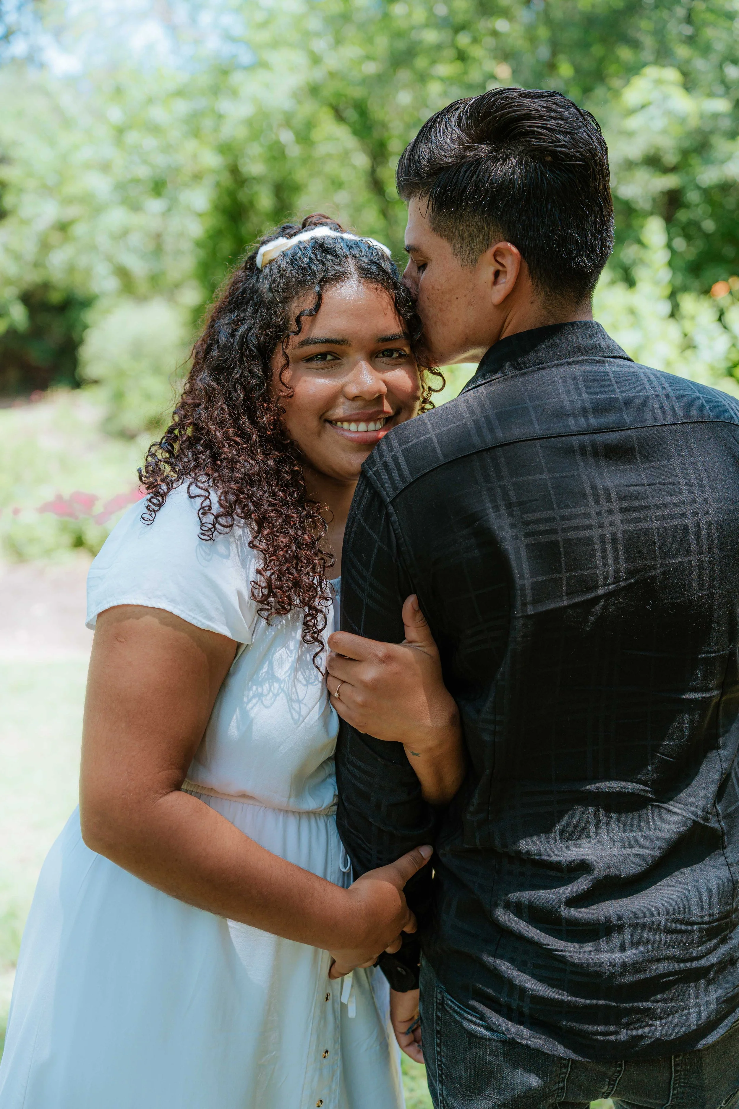 A couple embracing outdoors, with the man kissing the woman on her forehead. The woman is smiling at the camera, wearing a white dress, and the man is dressed in a black shirt with a checkered pattern. They are standing in a lush, green park or garde