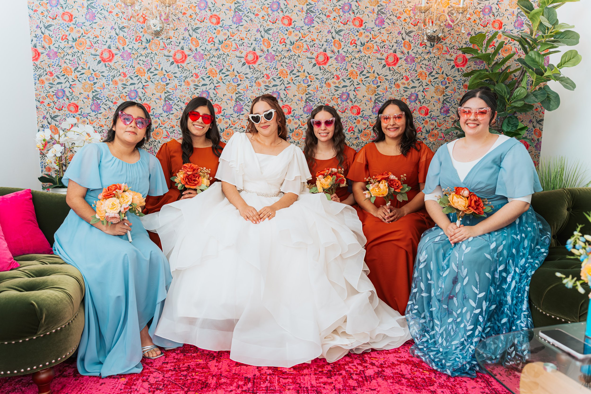 Group of women in colorful dresses and sunglasses sitting on a green couch, holding bouquets, with a floral wallpaper background and pink carpet.