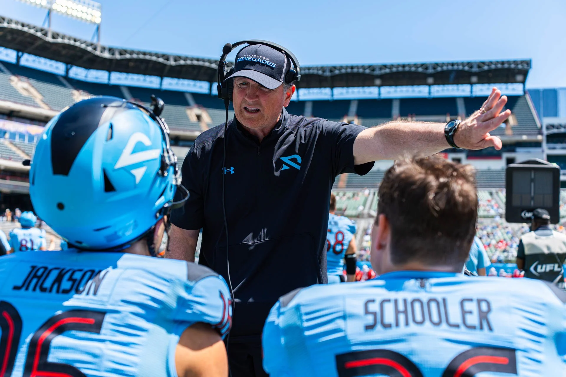 A coach wearing a black shirt with a blue logo, headset, and cap, giving instructions to two football players in light blue uniforms and helmets, on a football field at a stadium.