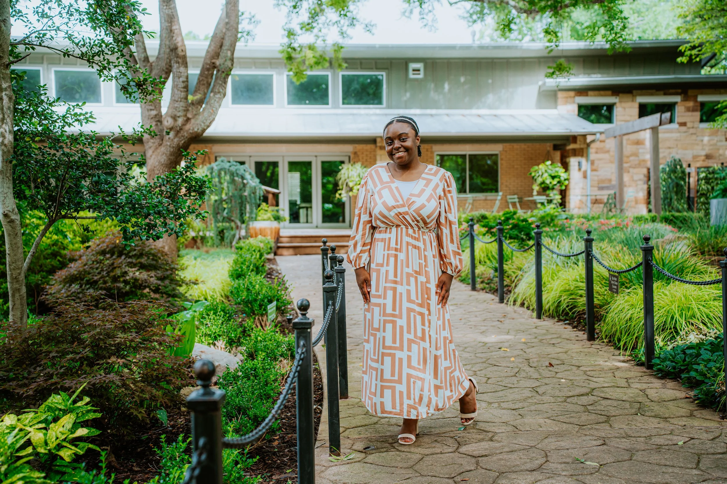 A woman in a white and beige geometric patterned dress standing on a paved garden pathway, smiling, with a house and greenery in the background.