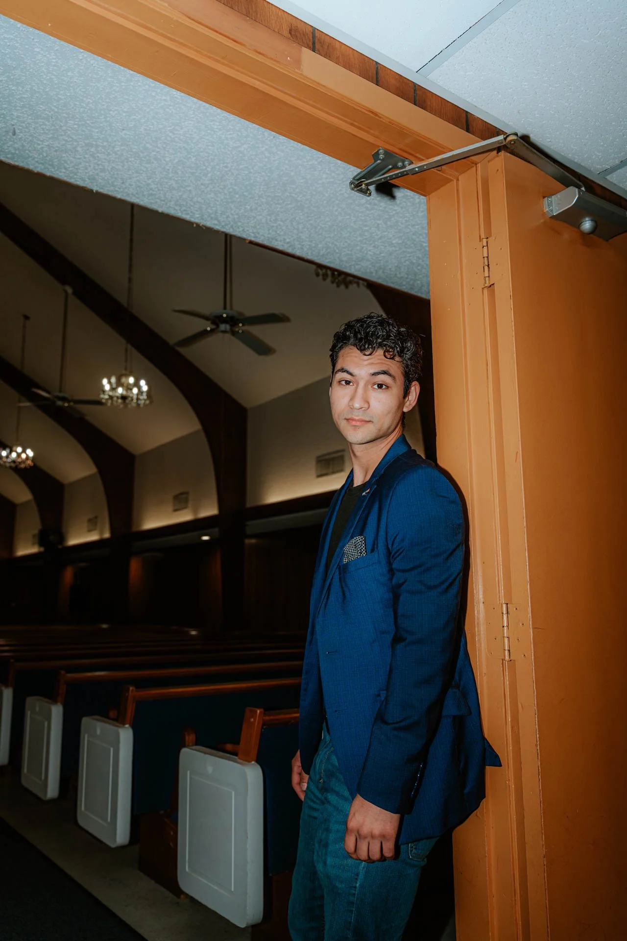 A young man in a blue blazer and jeans leaning against a doorframe in a church or auditorium with wooden beams and chandeliers.