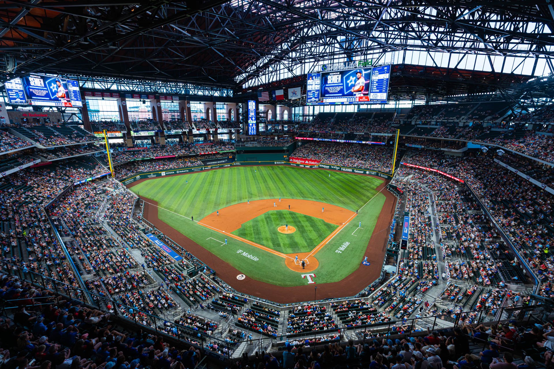 Interior of a baseball stadium filled with spectators, with players on the field preparing for a game. The field is green with dirt bases and a pitcher's mound, and large screens display player information above.