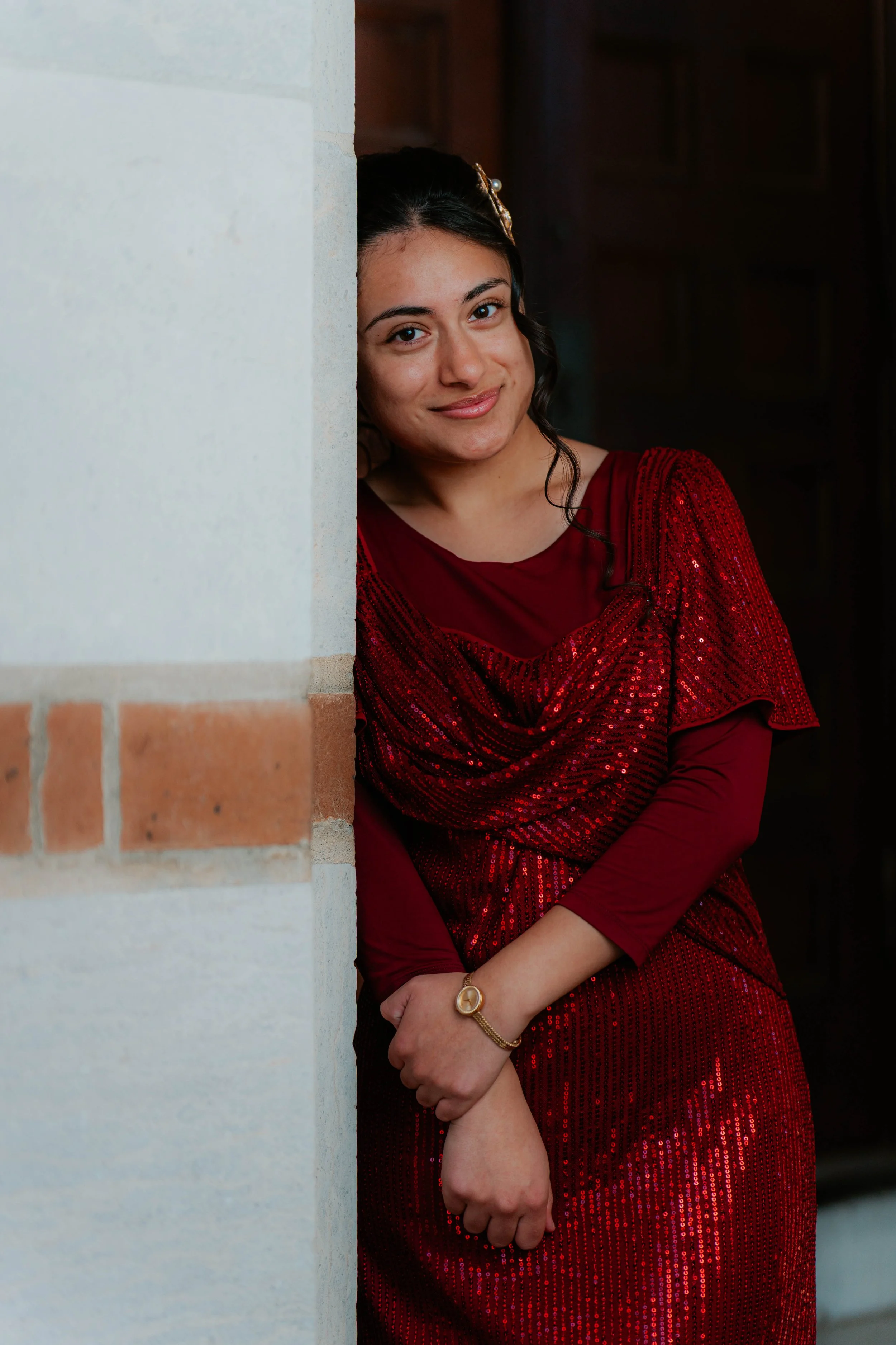 A woman in a red dress with sequins, smiling and peeking out from behind a wall.