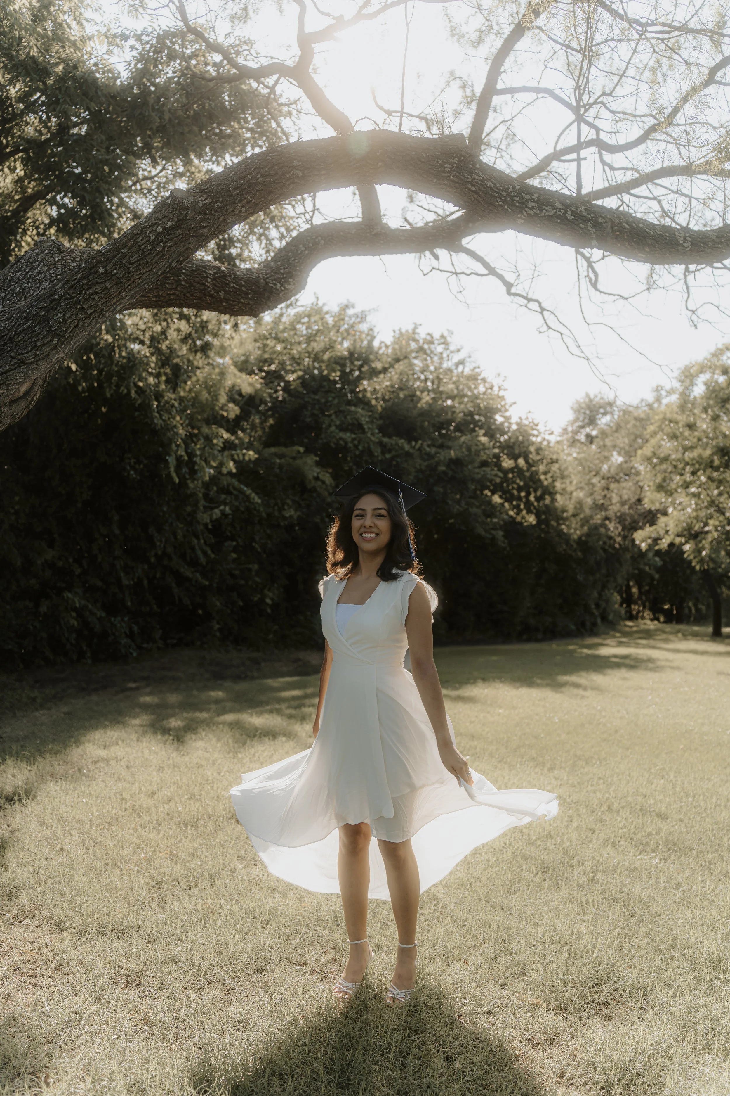 A woman in a white dress and graduation cap standing on a grassy field, smiling, with sunlight filtering through a large tree and foliage behind her.
