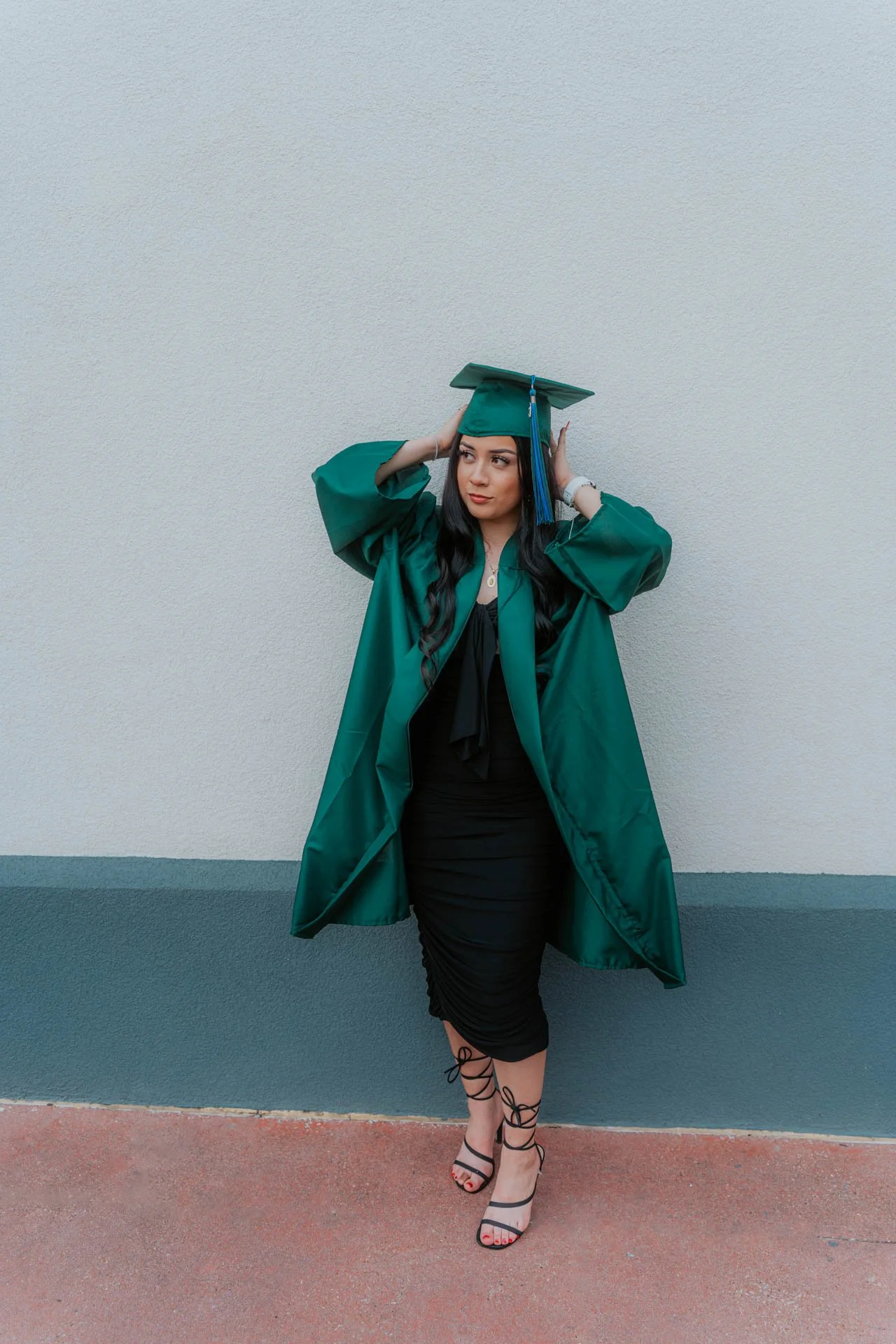 A woman in a green graduation gown and cap standing against a light-colored wall, adjusting her cap.