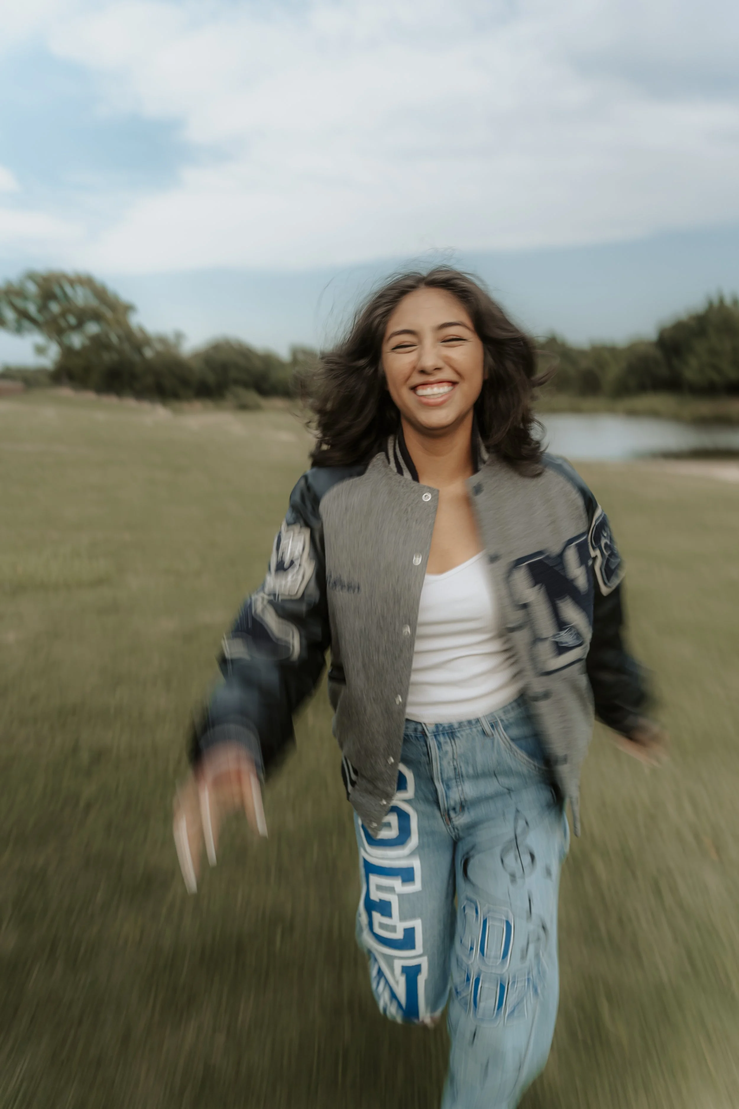 Young woman smiling and running in an open grassy field with trees and a small body of water under a partly cloudy sky.