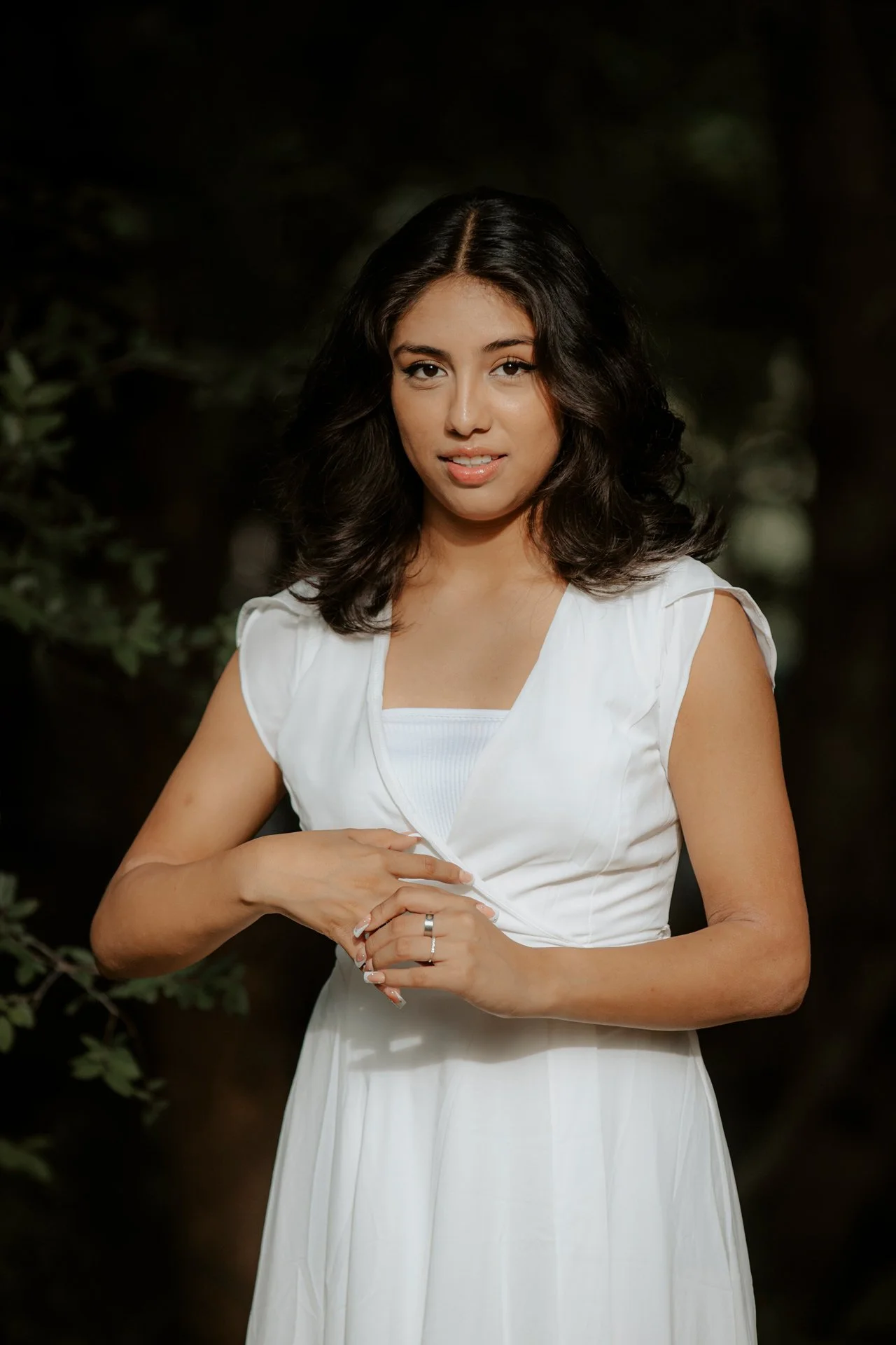 A woman in a white dress standing outdoors in a wooded area, with dark trees in the background.