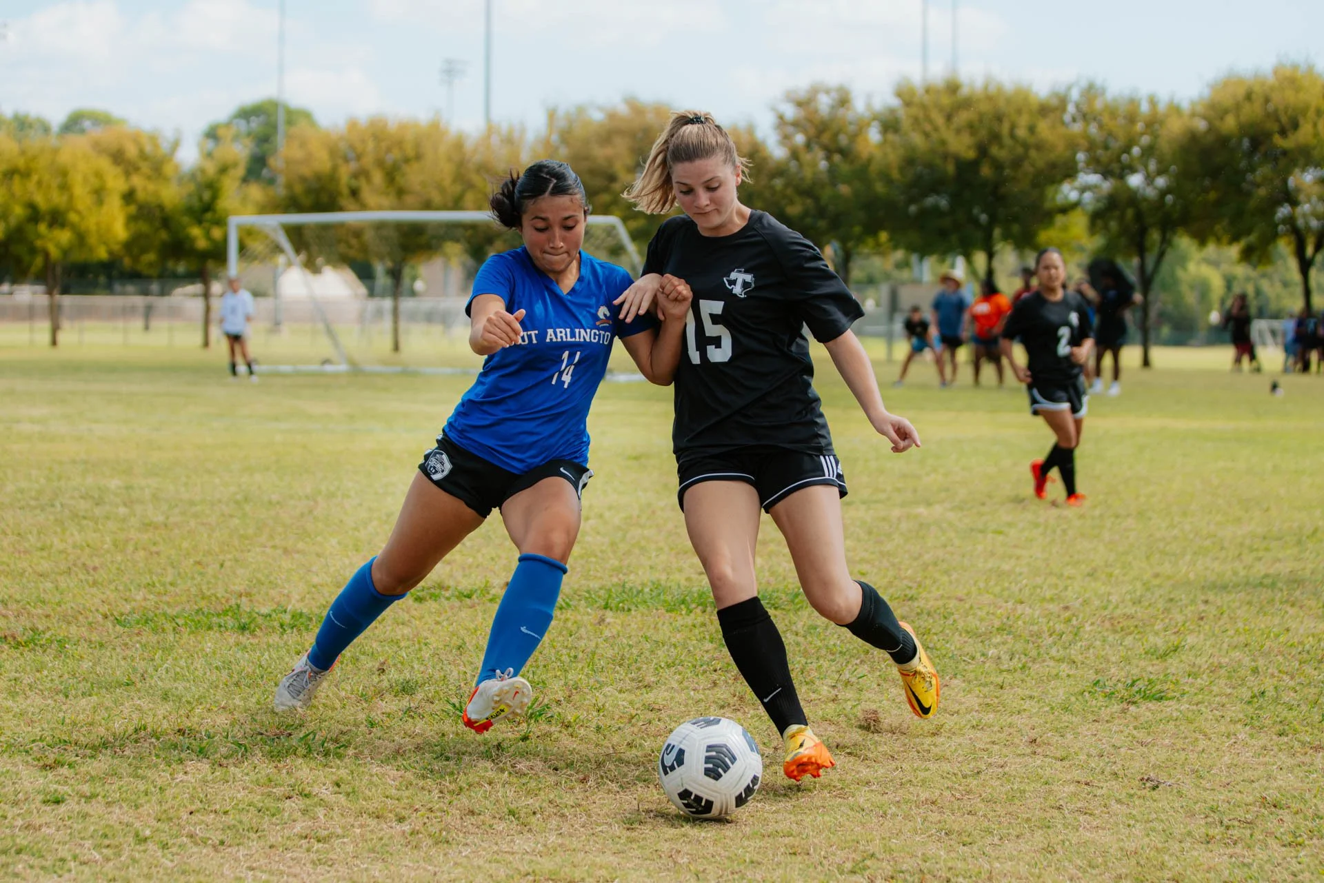 Two female soccer players, one in a blue jersey and the other in a black jersey, compete for the ball on a grassy field during a match, with trees and other players in the background.