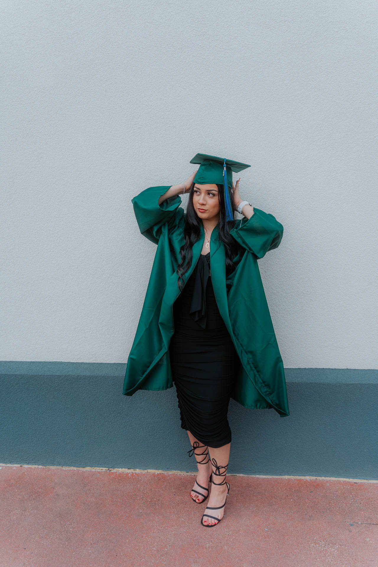 A young woman in a green graduate gown and cap standing against a gray textured wall, adjusting her cap, with black high-heeled sandals with lace-up straps.