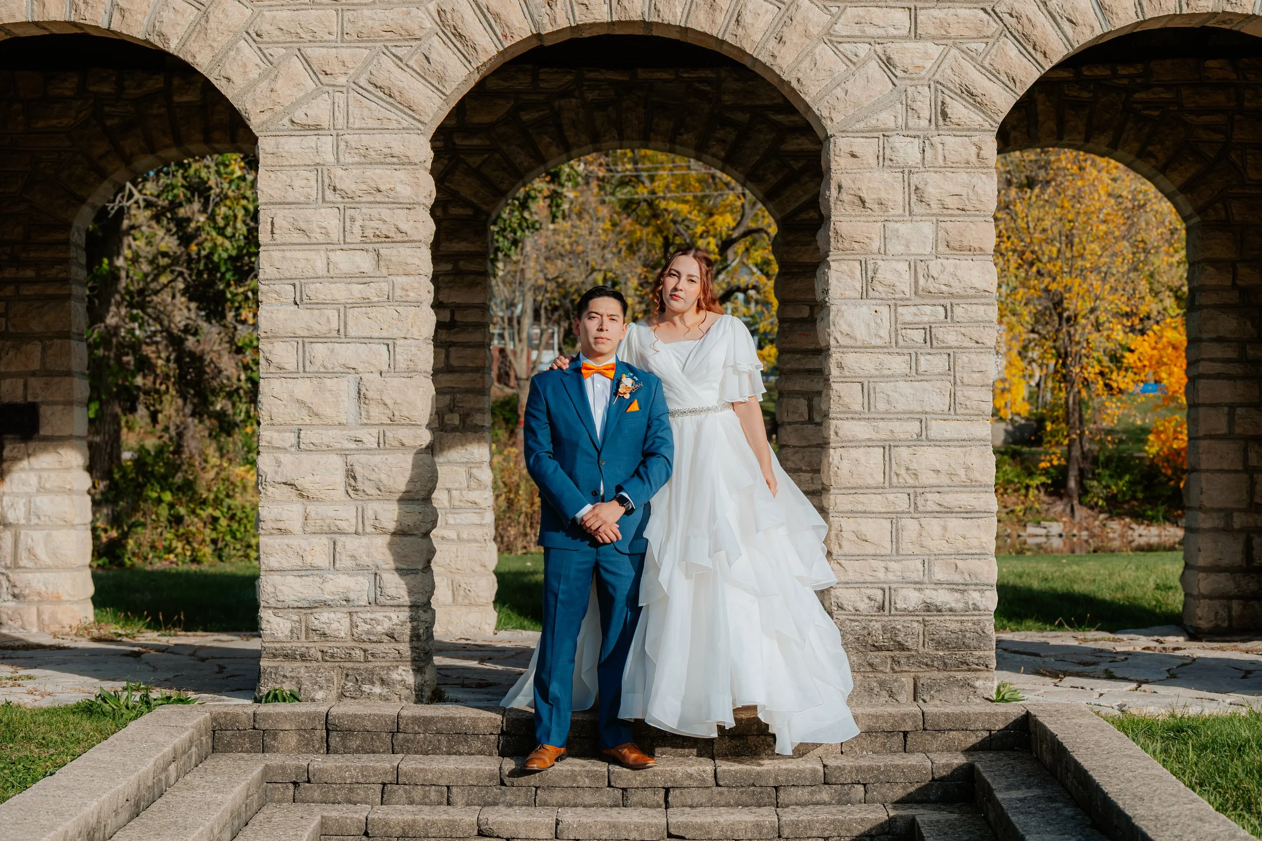 A wedding couple standing under stone arches outdoors during autumn. The groom is in a blue suit with a bow tie, and the bride is in a white wedding dress.
