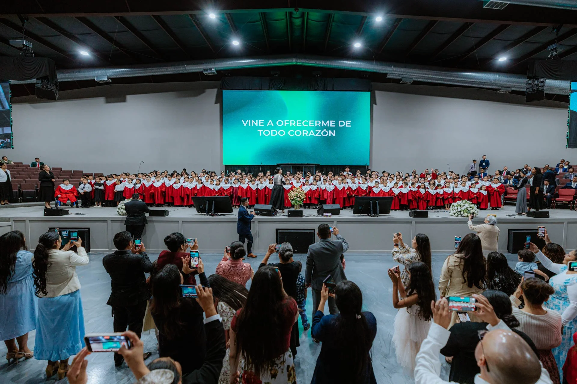 A large choir of children dressed in red robes and white collars performing on stage in a church or auditorium, with adults and audience members taking photos in the foreground, and a blue screen with Spanish text behind the choir that reads 'Vine a 