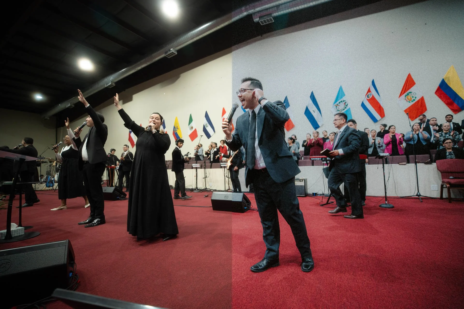A group of singers and musicians performing passionately on stage in a church or conference hall, with flags from various countries displayed on the wall behind them.