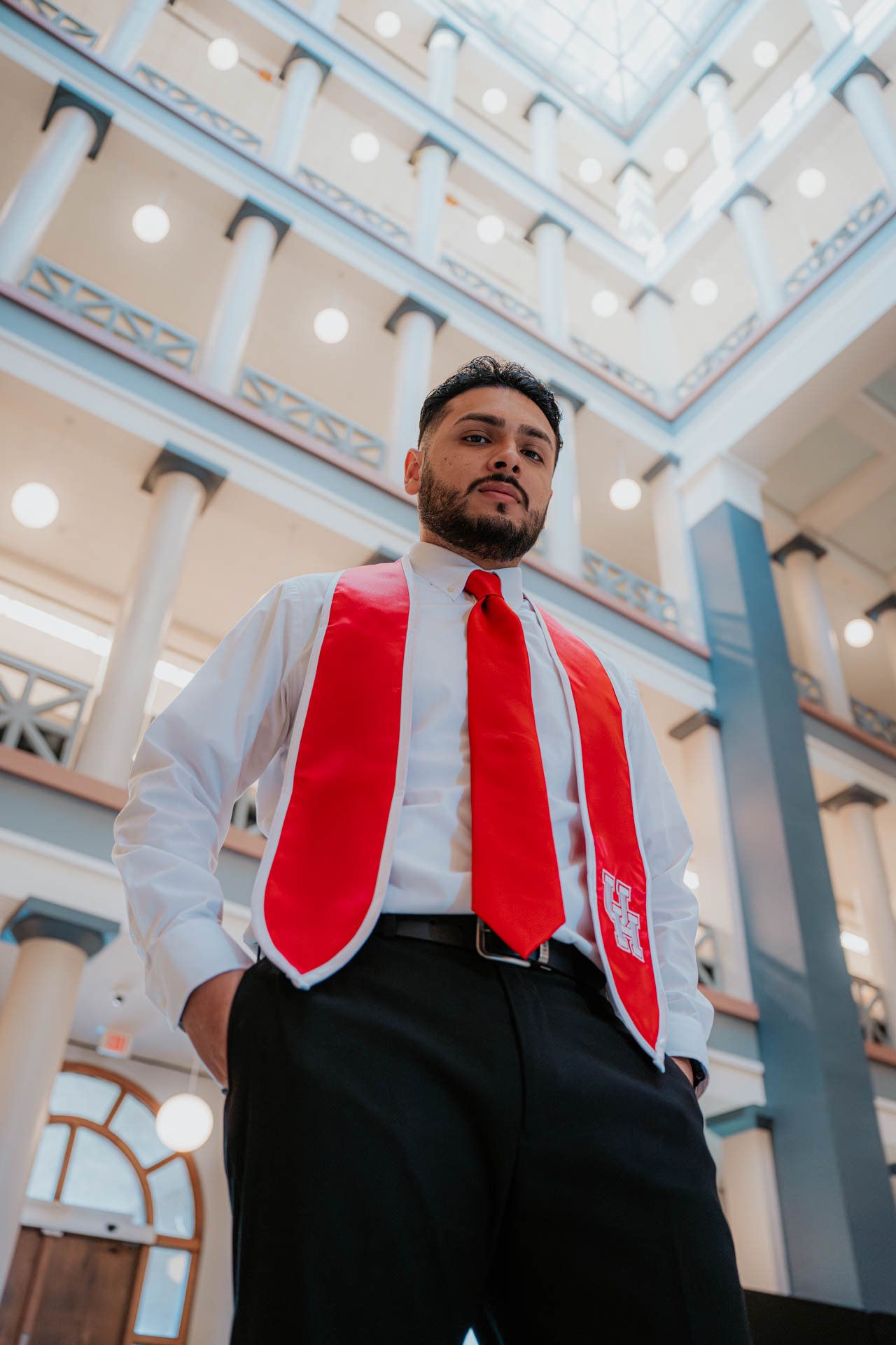 A man with a beard and black hair standing in a spacious, multi-story building with high ceilings, columns, and a glass ceiling, wearing a white shirt, red tie, and a red stole with a logo.