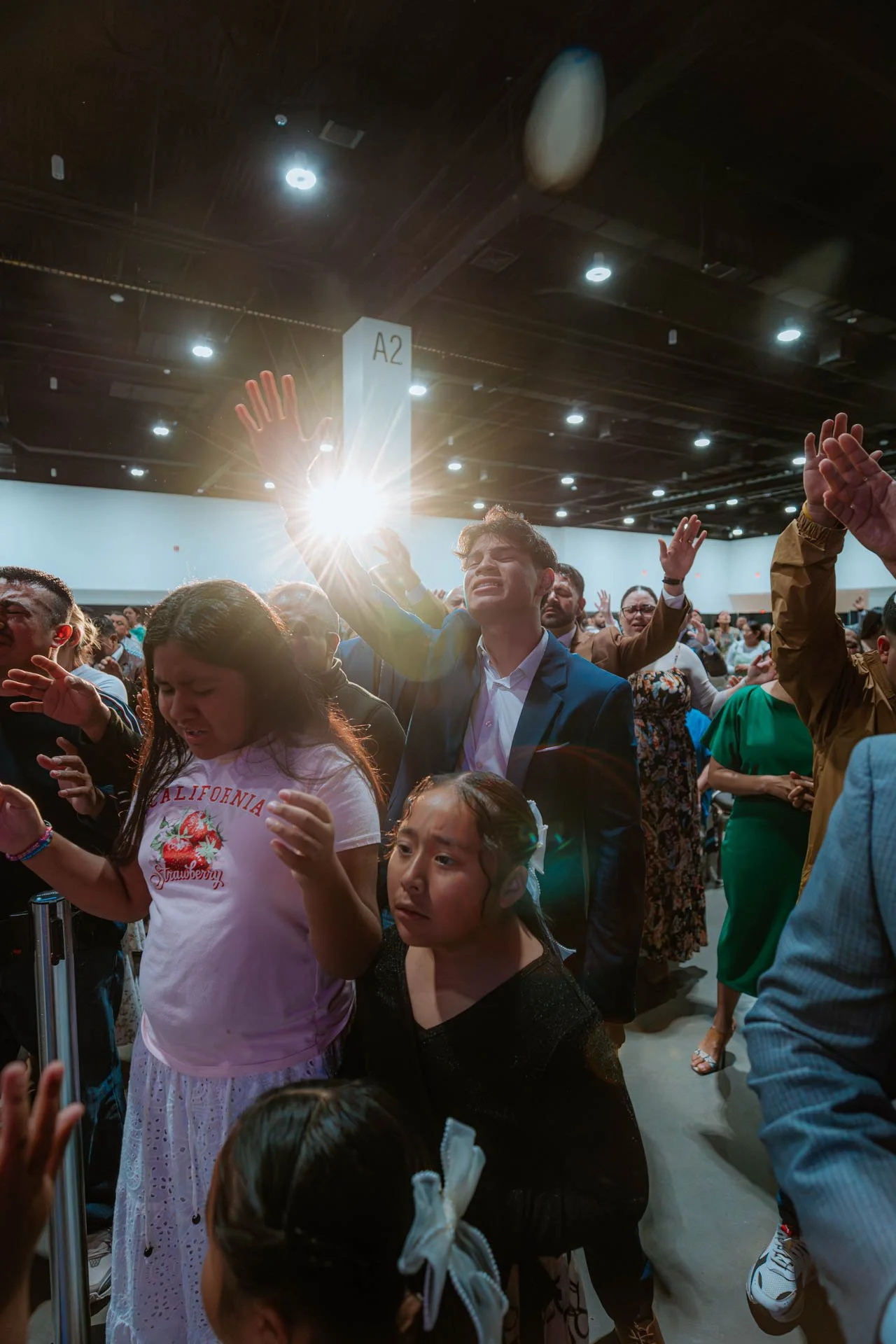 People at a gathering with raised hands, some eyes closed, in an indoor setting with overhead lights and a sign labeled A2.